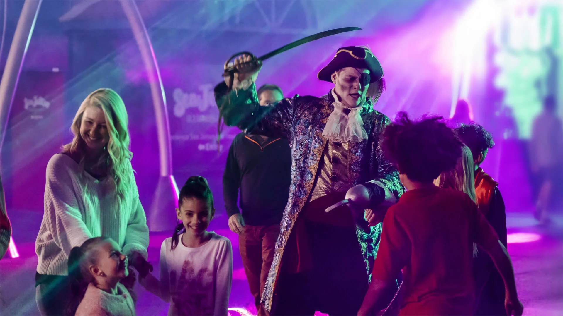 A person dressed as a pirate with a sword entertains a group of smiling children and a woman under colorful, vibrant lights at night.