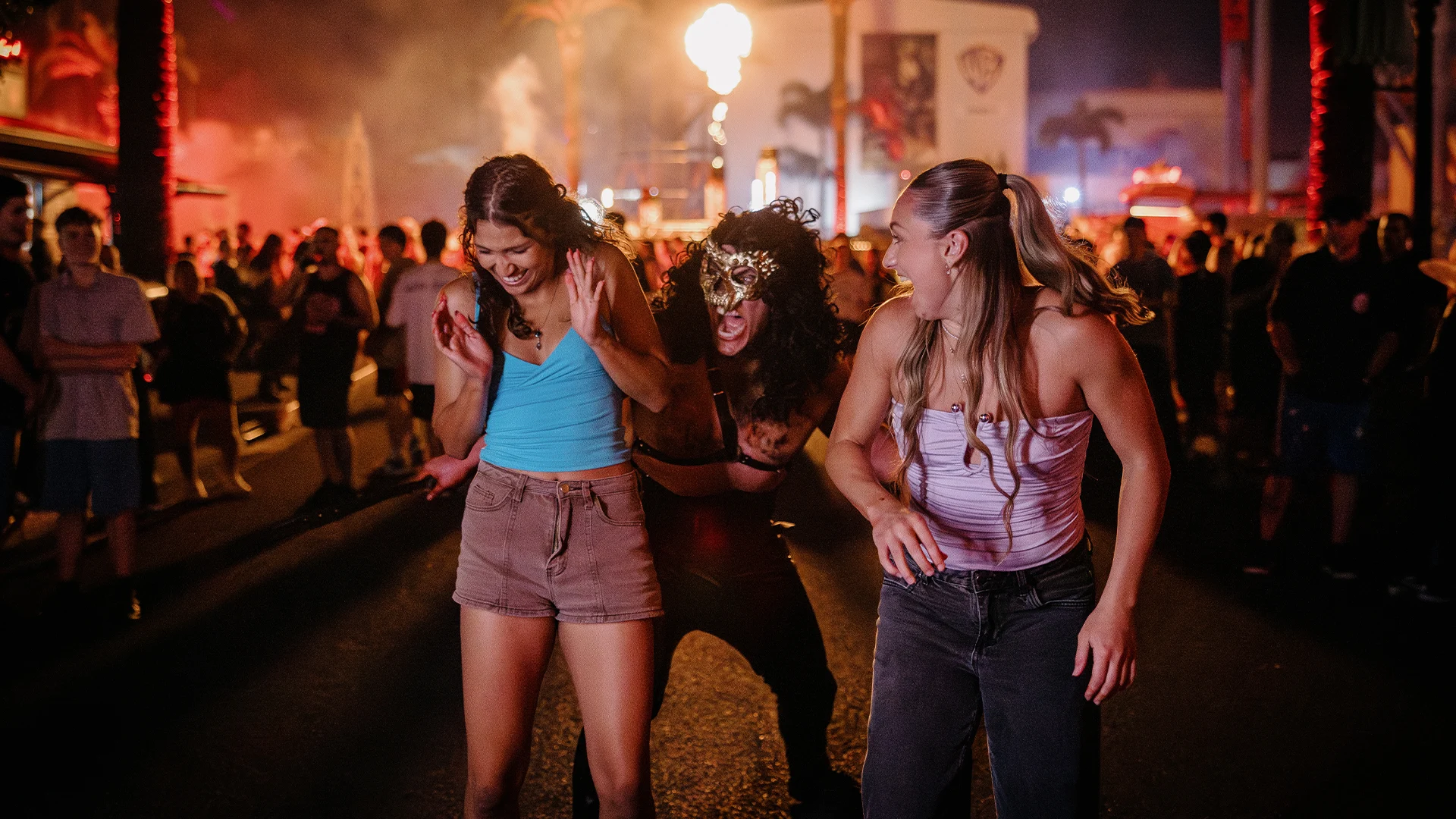 Three women react with surprise and laughter as a person in a scary mask sneaks up behind them at a lively, nighttime outdoor event with fire and crowds in the background.