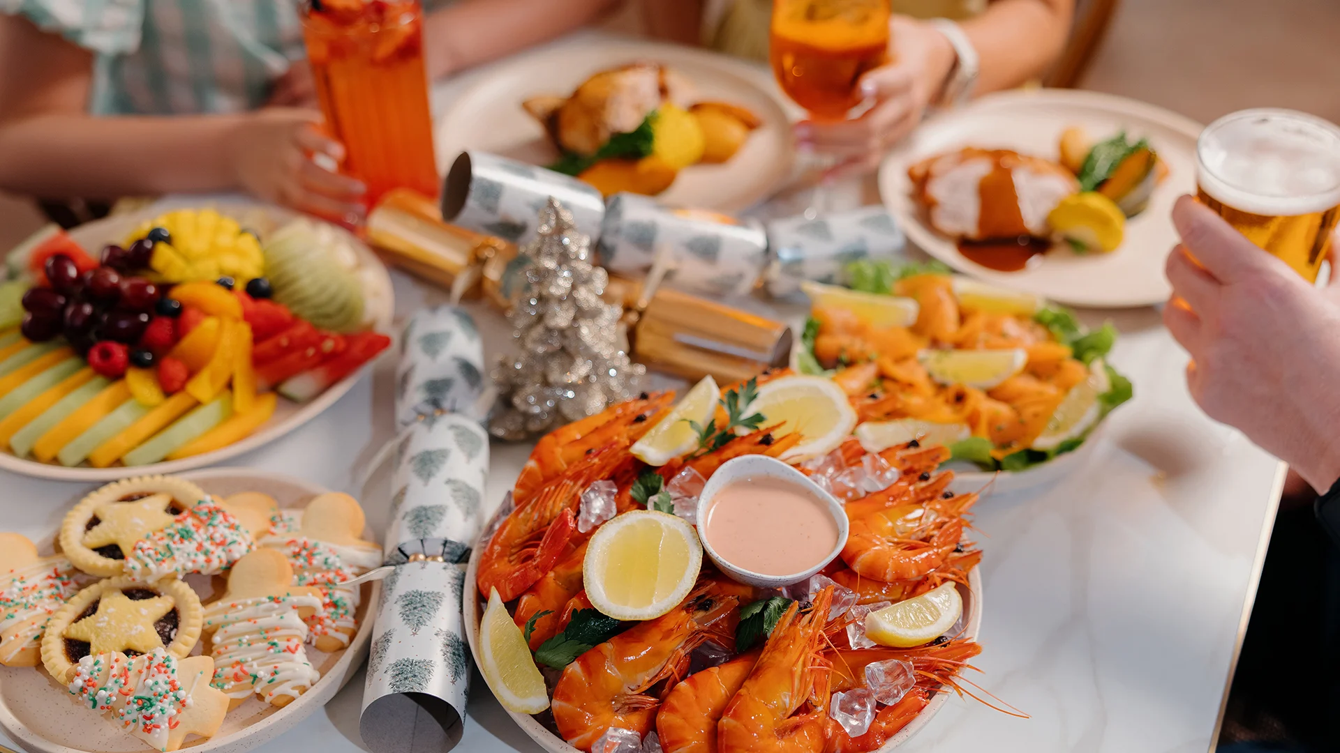 A festive table set with plates of prawns, lemon wedges, fruit, cookies, and meat dishes. Three people hold drinks, and holiday decorations, including a silver tree, add a celebratory touch.