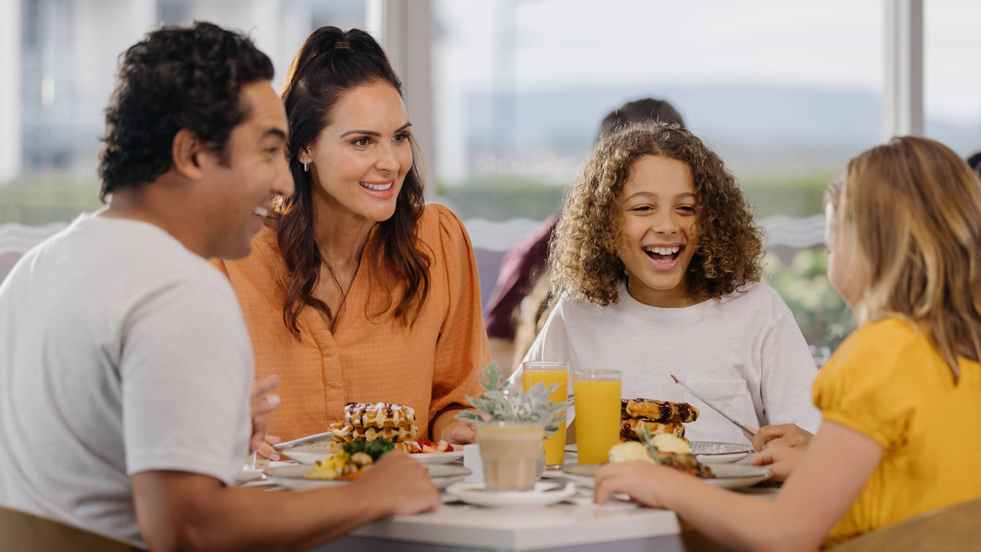A family of four sits at a dining table at Shoreline Restaurant, smiling and enjoying a buffet breakfast meal together, with plates of food and drinks visible on the table.