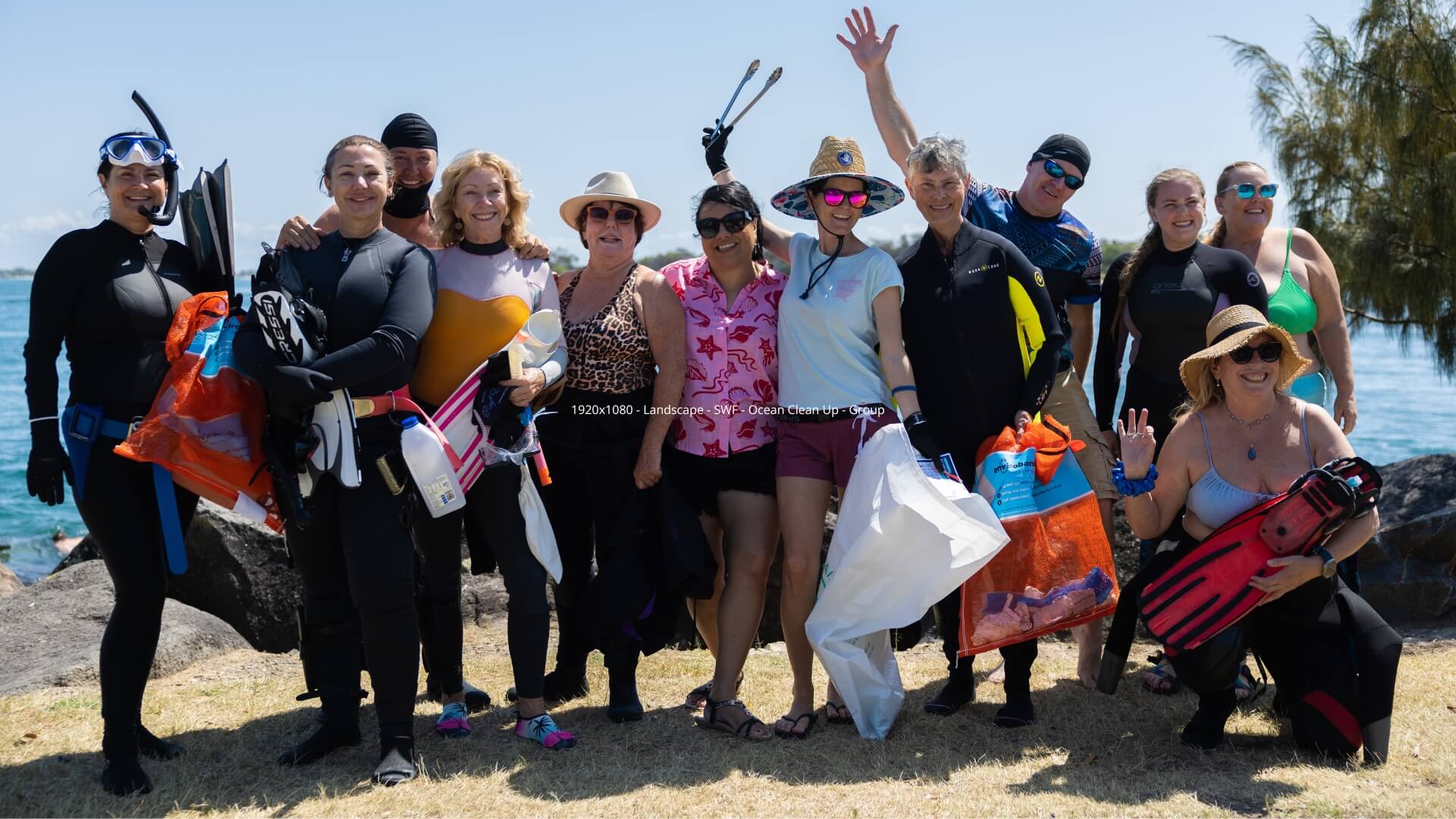 A group of people in swim and snorkel gear stand outdoors near water, smiling and holding bags of collected trash.