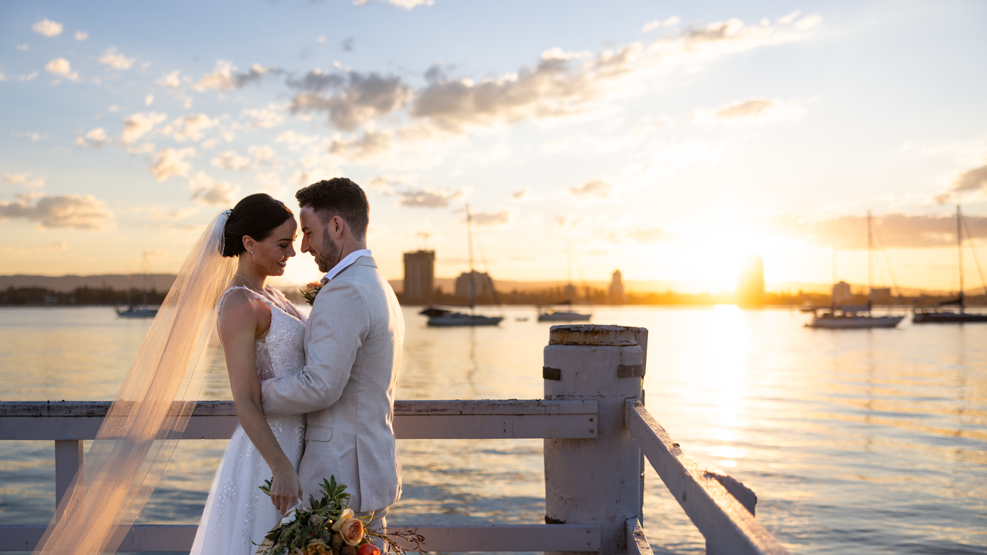 A bride and groom stand closely together on a pier at sunset, with sailboats and a city skyline in the background. The bride holds a bouquet and wears a veil, while the groom is dressed in a light suit.