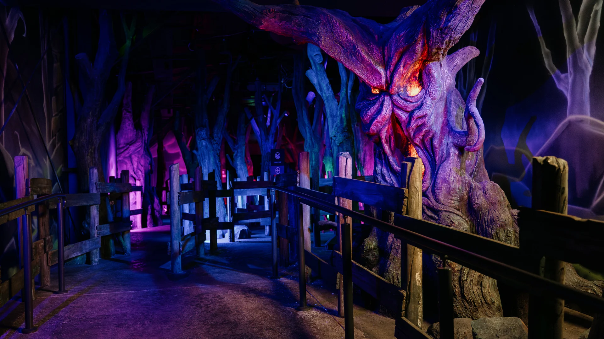 A dimly lit, eerie indoor forest scene with twisted trees, including one with a monstrous face glowing orange and purple, surrounded by wooden railings and surreal lighting effects.