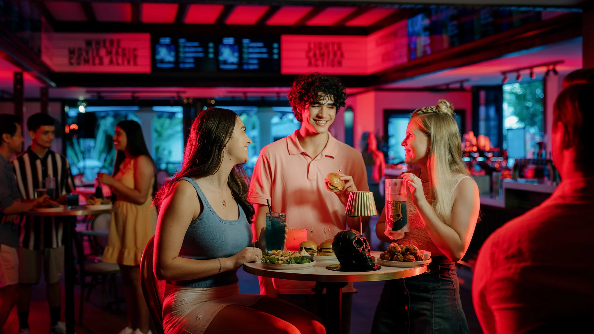 Three young adults laugh and chat around a high table with drinks and snacks in a colorful, modern bar or café with neon lighting. Other patrons and menu boards are visible in the background.
