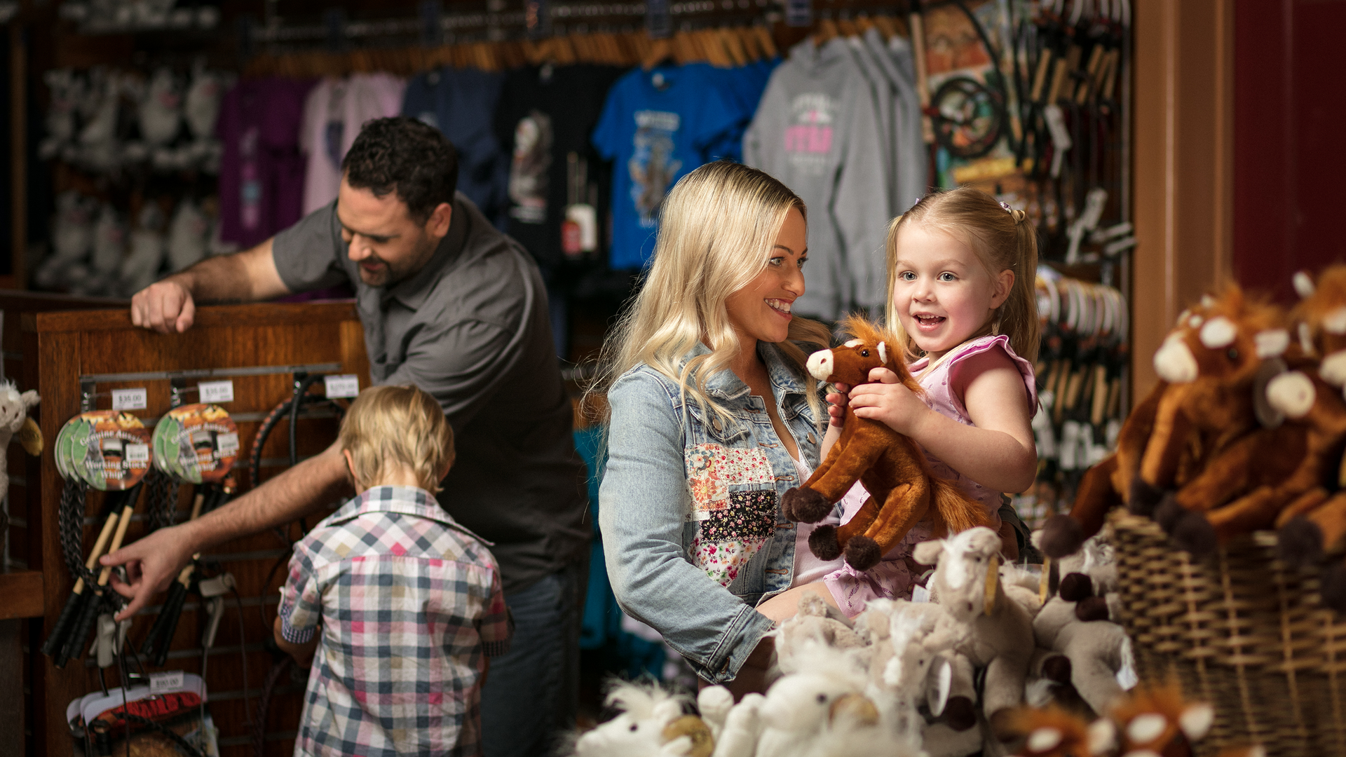 A smiling woman stands with two children in a shop filled with plush toy horses and souvenir shirts; one child hugs a stuffed horse, while a man browses merchandise in the background.