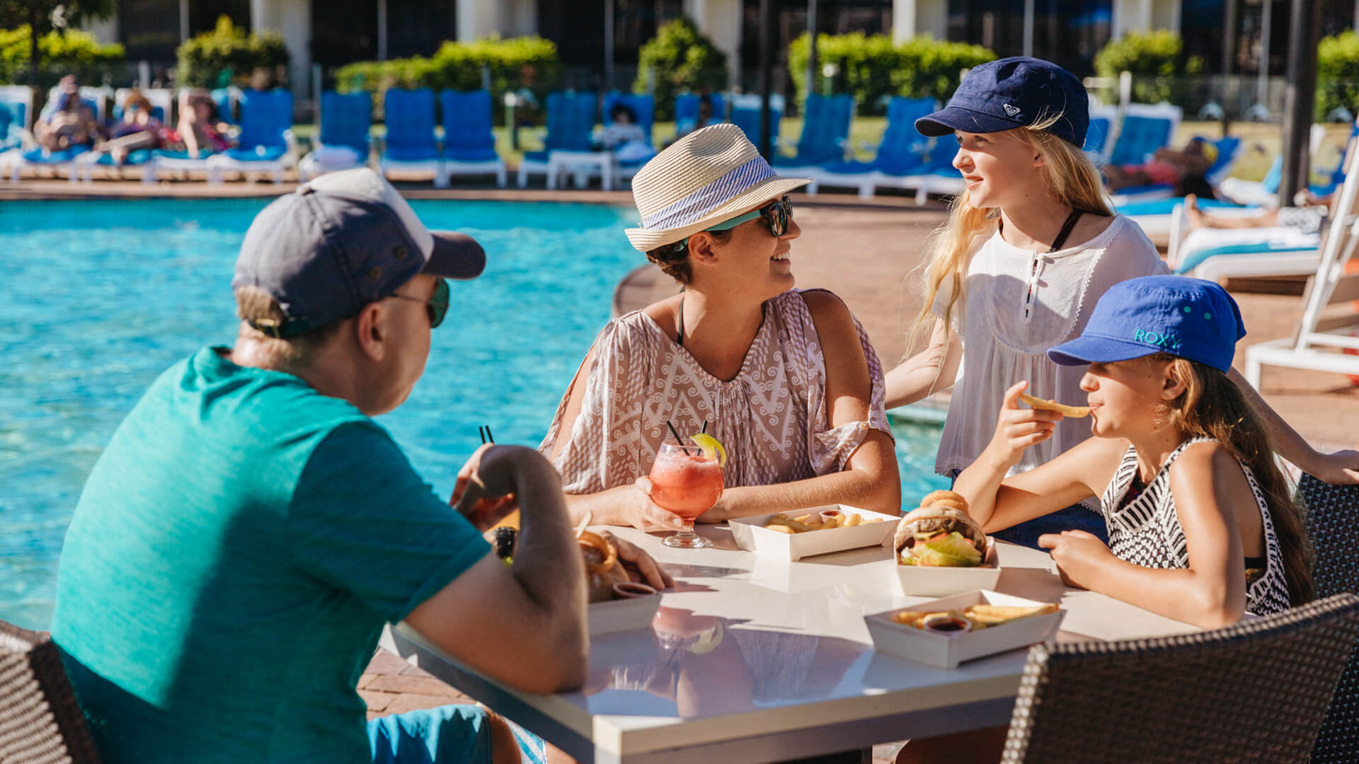 Family of four enjoying a meal and drinks at an outdoor table by the pool at Sea World Resort, with blue loungers and greenery in the background.