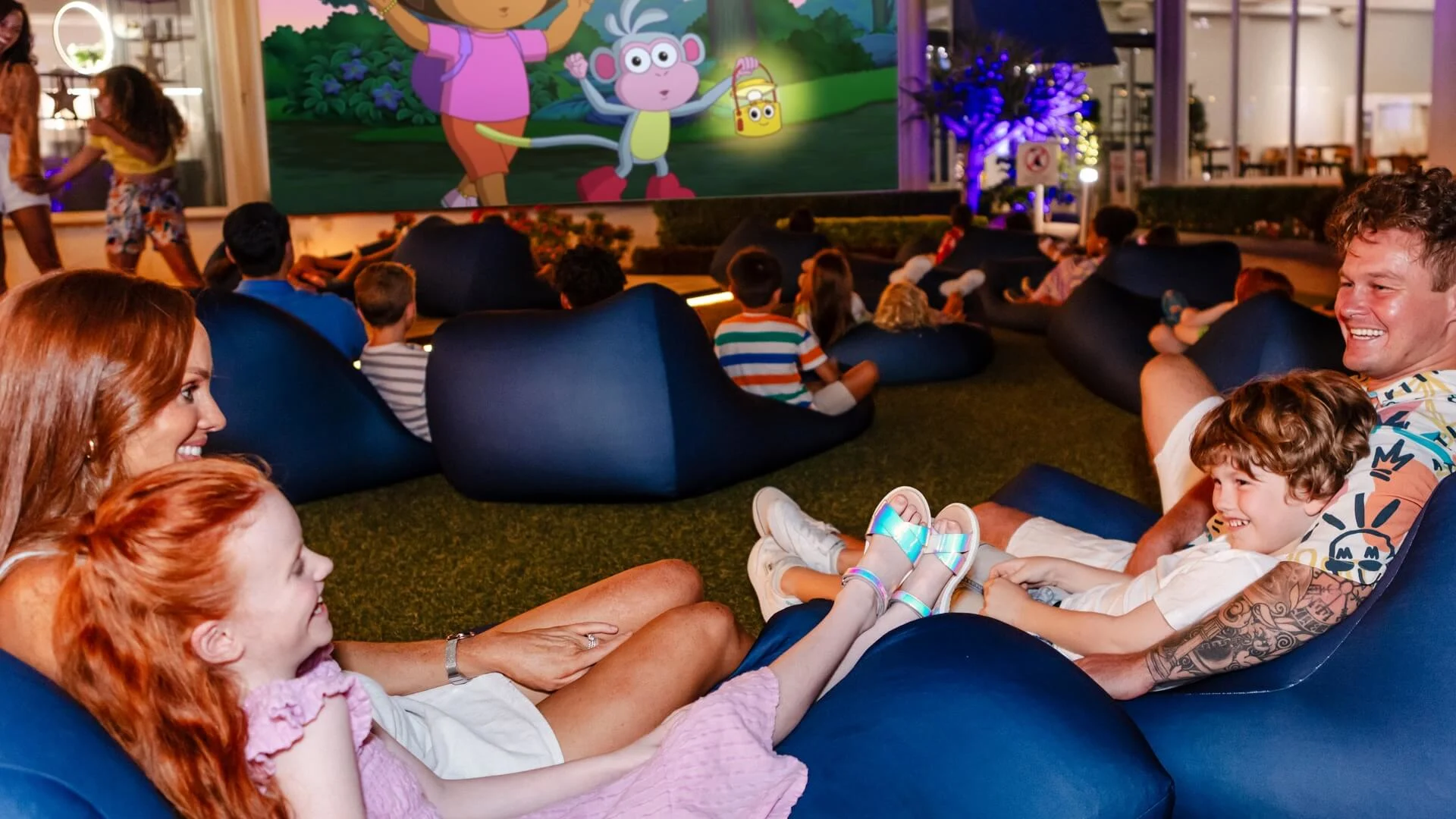 A mother and daughter, as well as families in the background, watch a Dora the Explorer movie on a big screen.