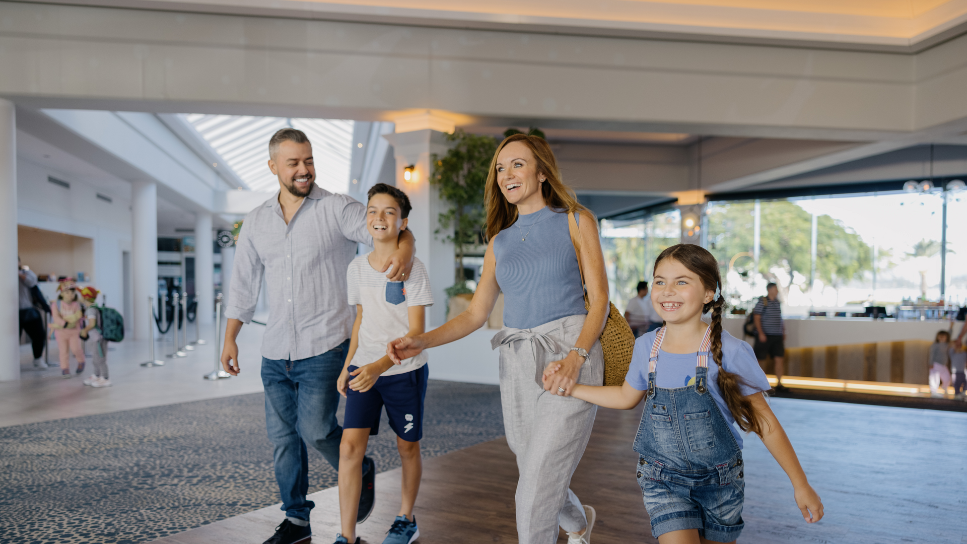 A family of four walks through a bright indoor space. The parents hold hands with their two children, who are smiling.