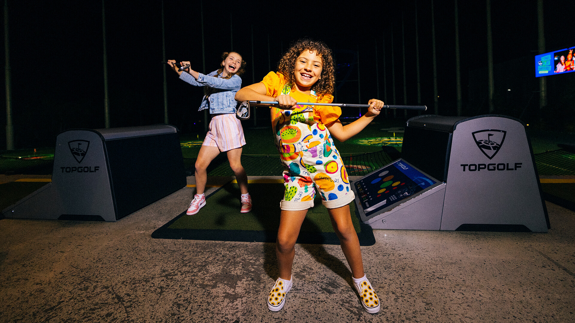 Two children smiling and posing with golf clubs at an indoor golf venue, with Topgolf branding visible in the background.