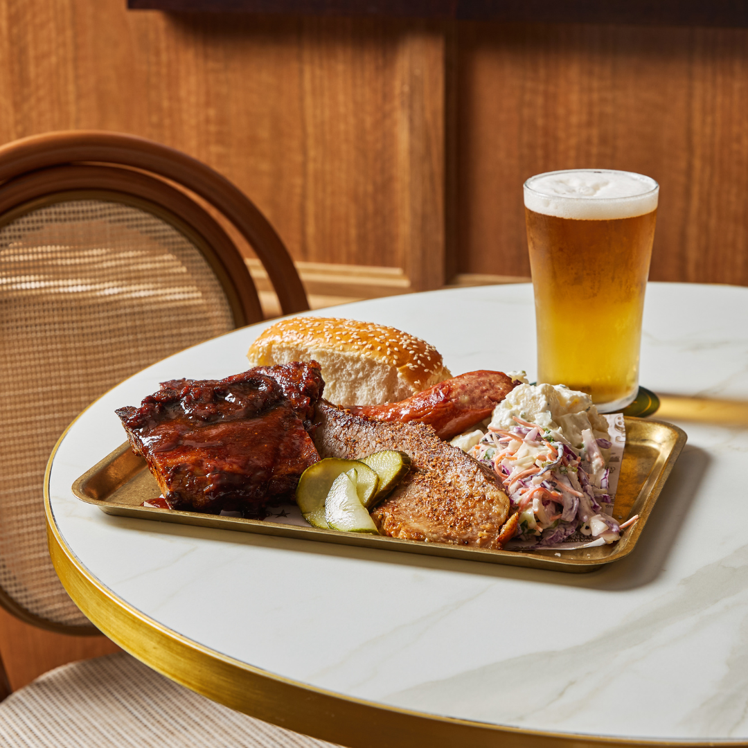 A plated meal with barbecue ribs, sausage, coleslaw, pickles, cornbread, and a sesame bun sits on a round marble table next to a glass of beer and a wooden chair.