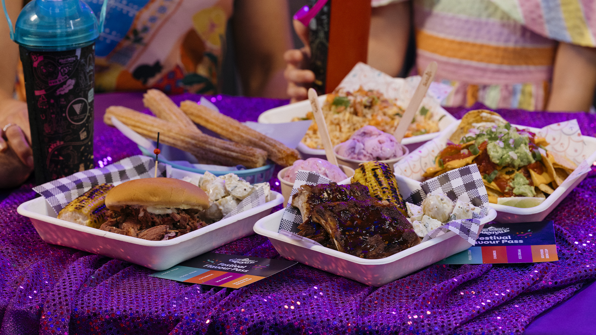 A table covered with a purple sequin cloth displays various festival foods, including churros, BBQ ribs, a pulled pork sandwich, seasoned fries, grilled corn, coleslaw, ice cream, and a souvenir drink cup.