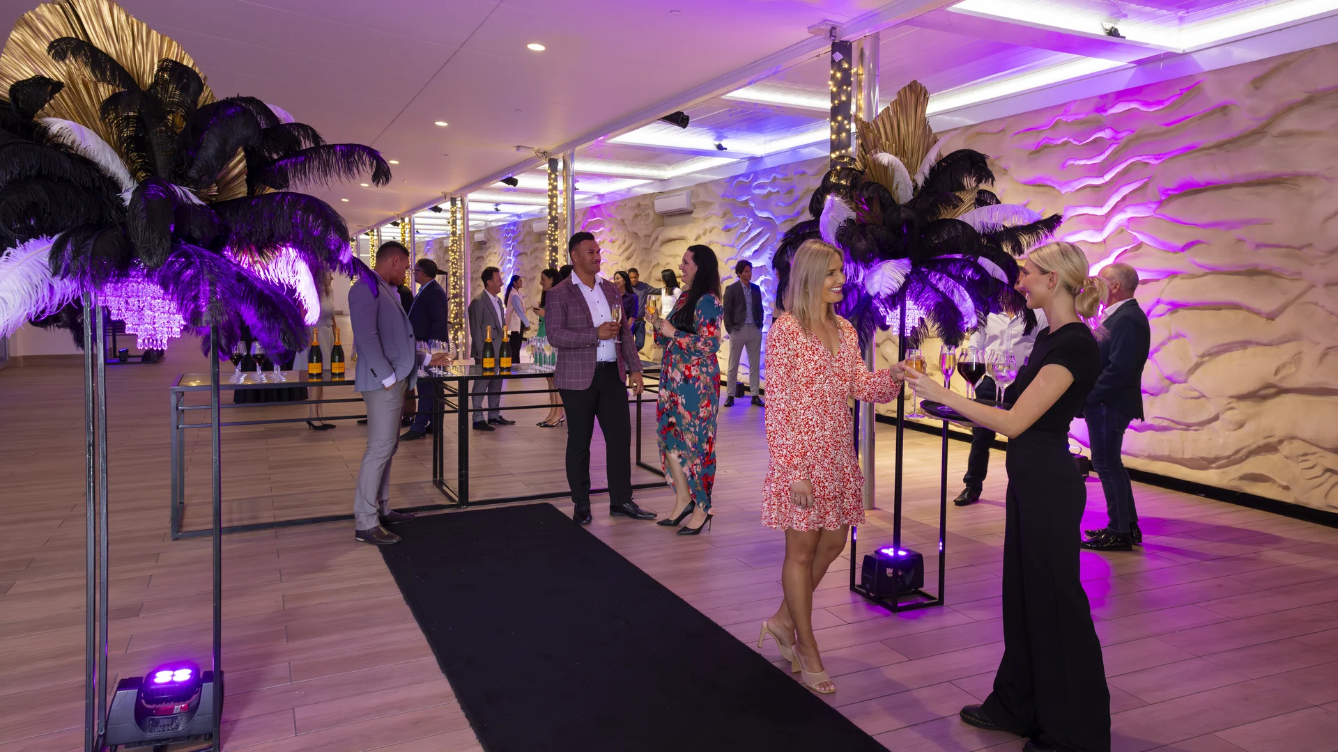 People in formal attire mingle at a stylish indoor event with purple lighting, decorative black and gold feather arrangements, and drinks displayed on a table in the background.