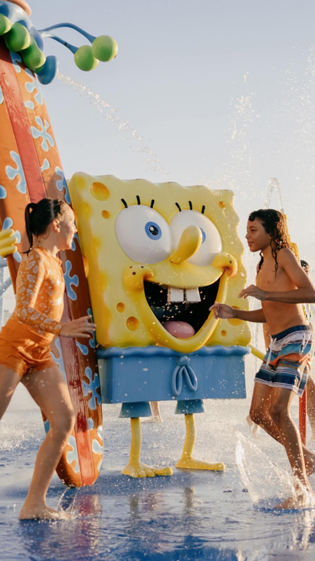 Two children in swimsuits play near a large SpongeBob SquarePants character at a water park, with water spraying around them.