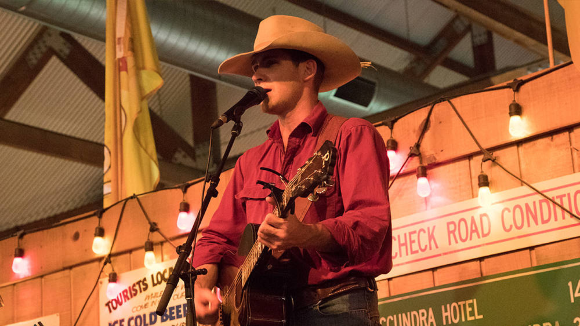 A man in a cowboy hat and red shirt plays an acoustic guitar and sings into a microphone on stage, with colorful string lights and vintage signs in the background.