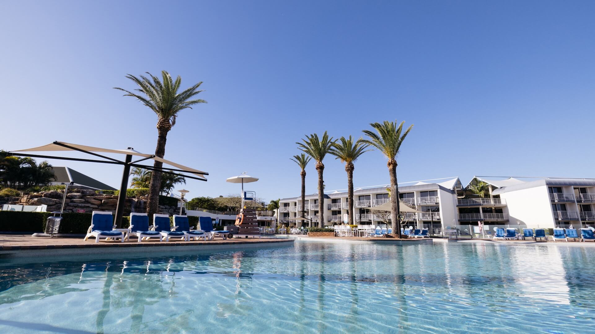 Outdoor swimming pool with clear water, surrounded by lounge chairs and palm trees. Two-story white buildings in the background under a clear blue sky.