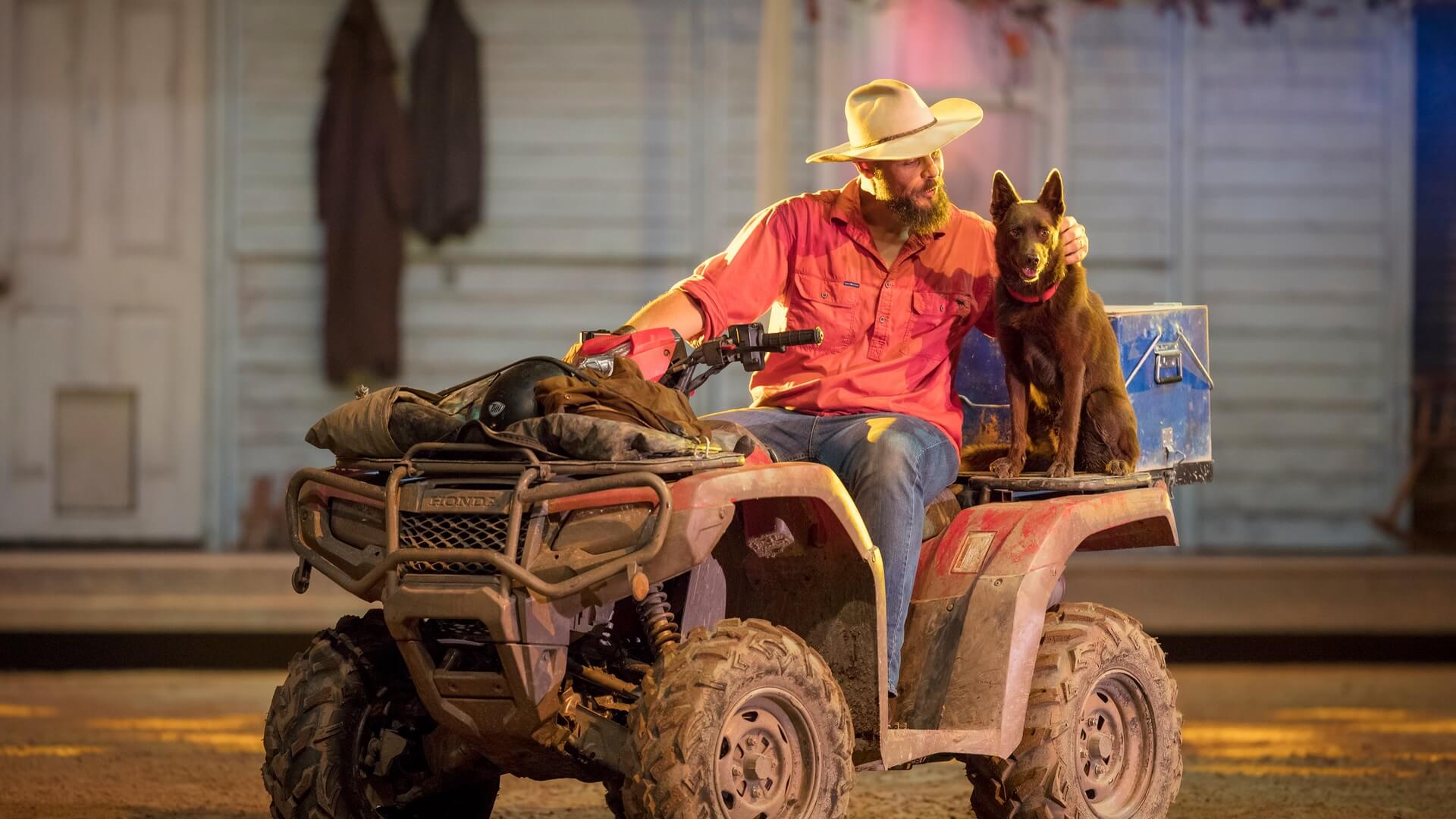 Stockman in an Australian Outback Spectacular show sitting on a quad bike, sharing a moment with his working dog.