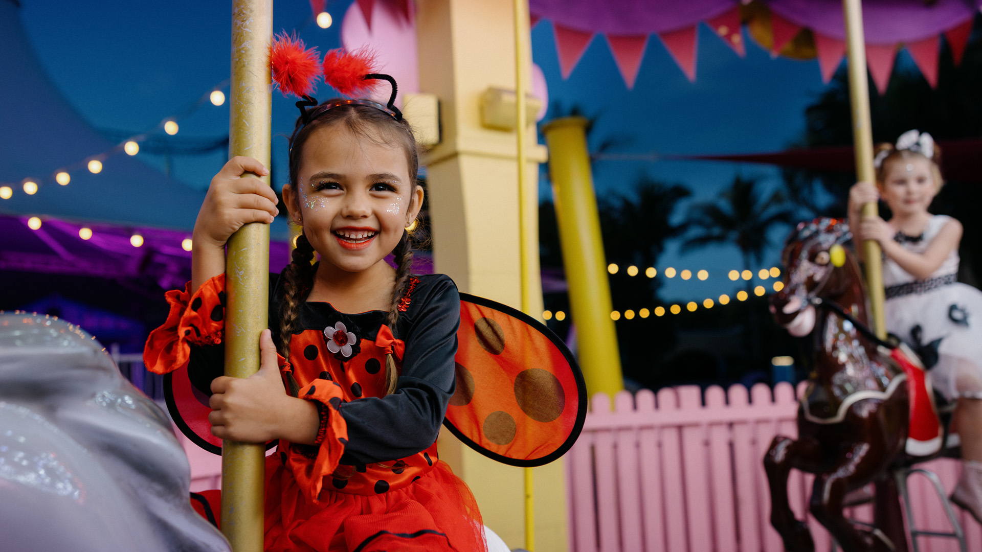A young girl in a ladybug costume smiles while riding a carousel horse at an amusement park, with another child and festive lights visible in the background.