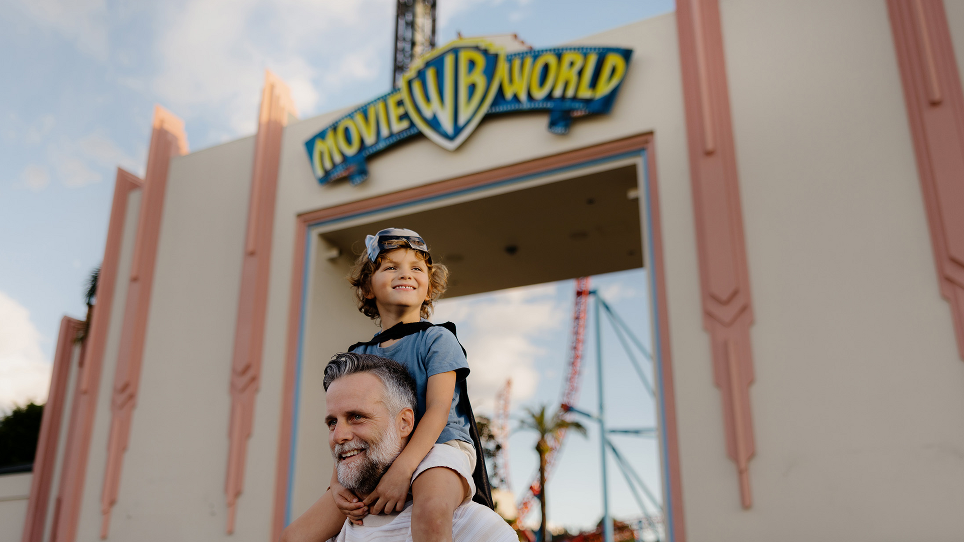 A smiling man carries a happy child on his shoulders in front of the entrance gate to Movie World theme park, with the park's blue and yellow sign visible above them.