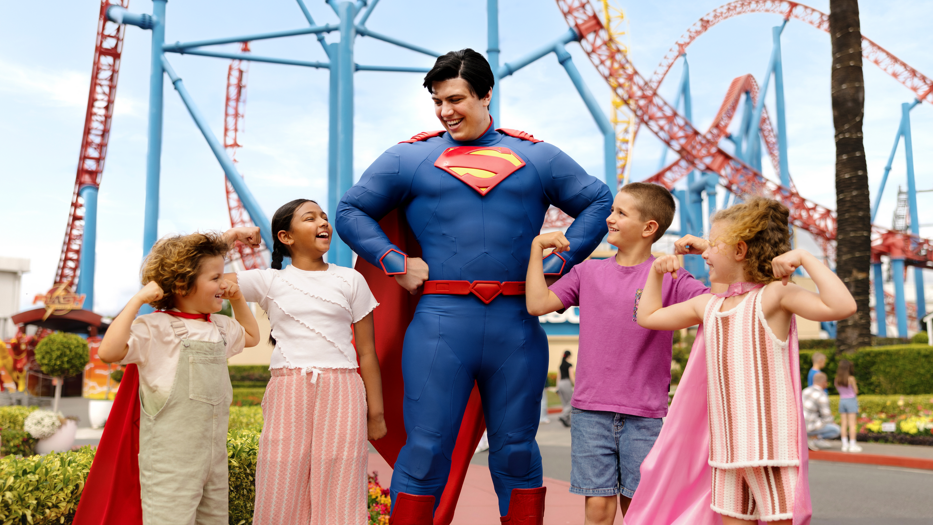 A person dressed as Superman poses with four smiling children at an amusement park, with roller coasters in the background. The children are flexing their arms and wearing capes, appearing excited and playful.