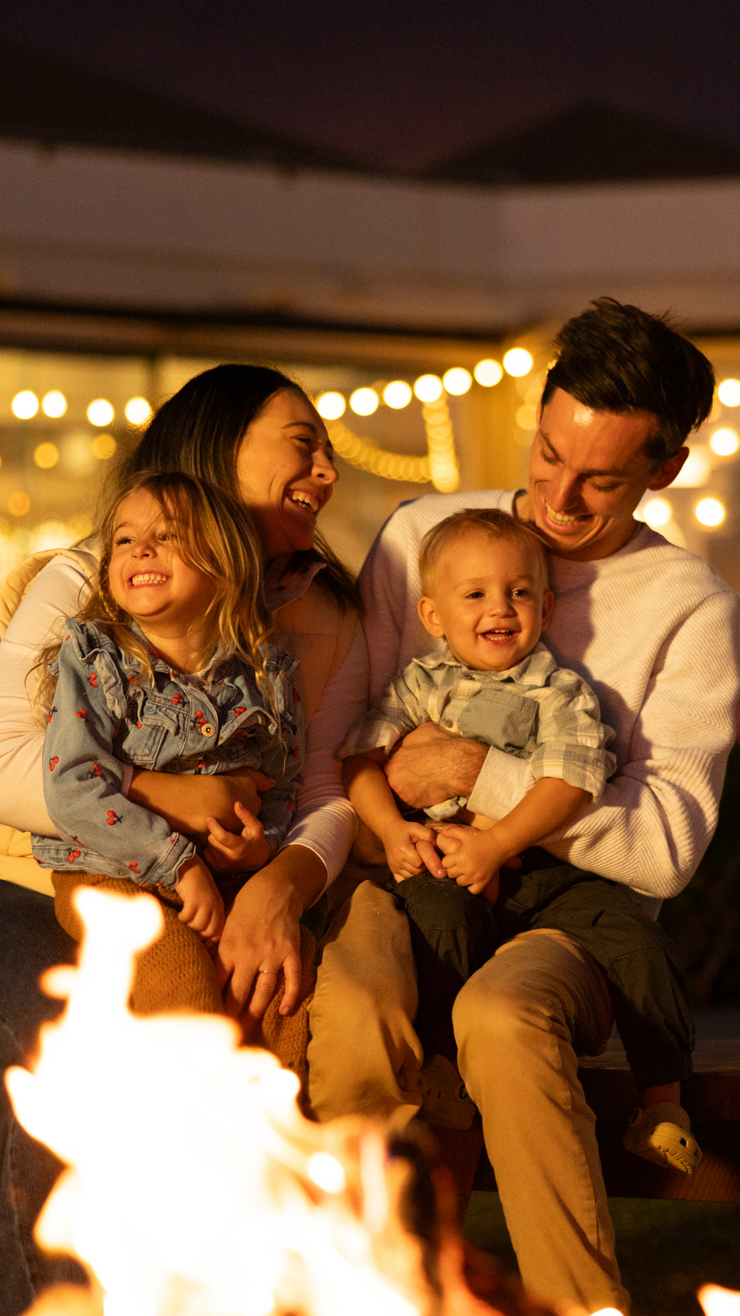 A smiling family of four sits on a bench by a fire pit at night, with warm string lights and a building in the background. The parents hold their young children, all appearing happy and relaxed.