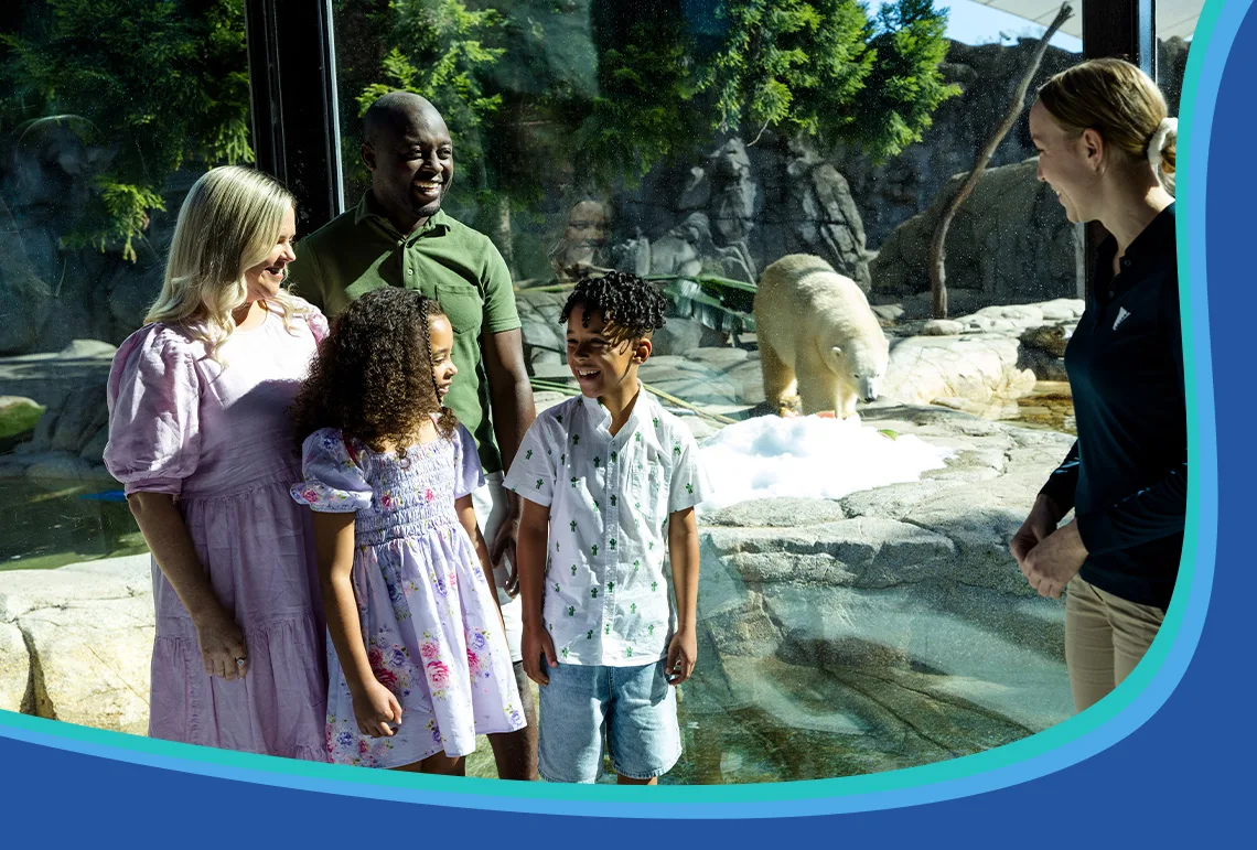 A family with two children stands by a glass enclosure, smiling and talking with a zoo staff member; a polar bear is visible in the background.
