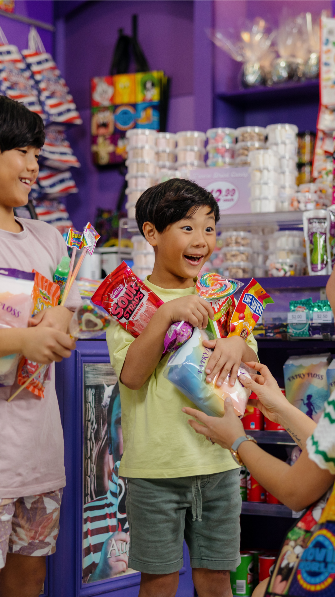 A smiling child holds an assortment of candy while another child and an adult hand over more treats in a colorful candy store.