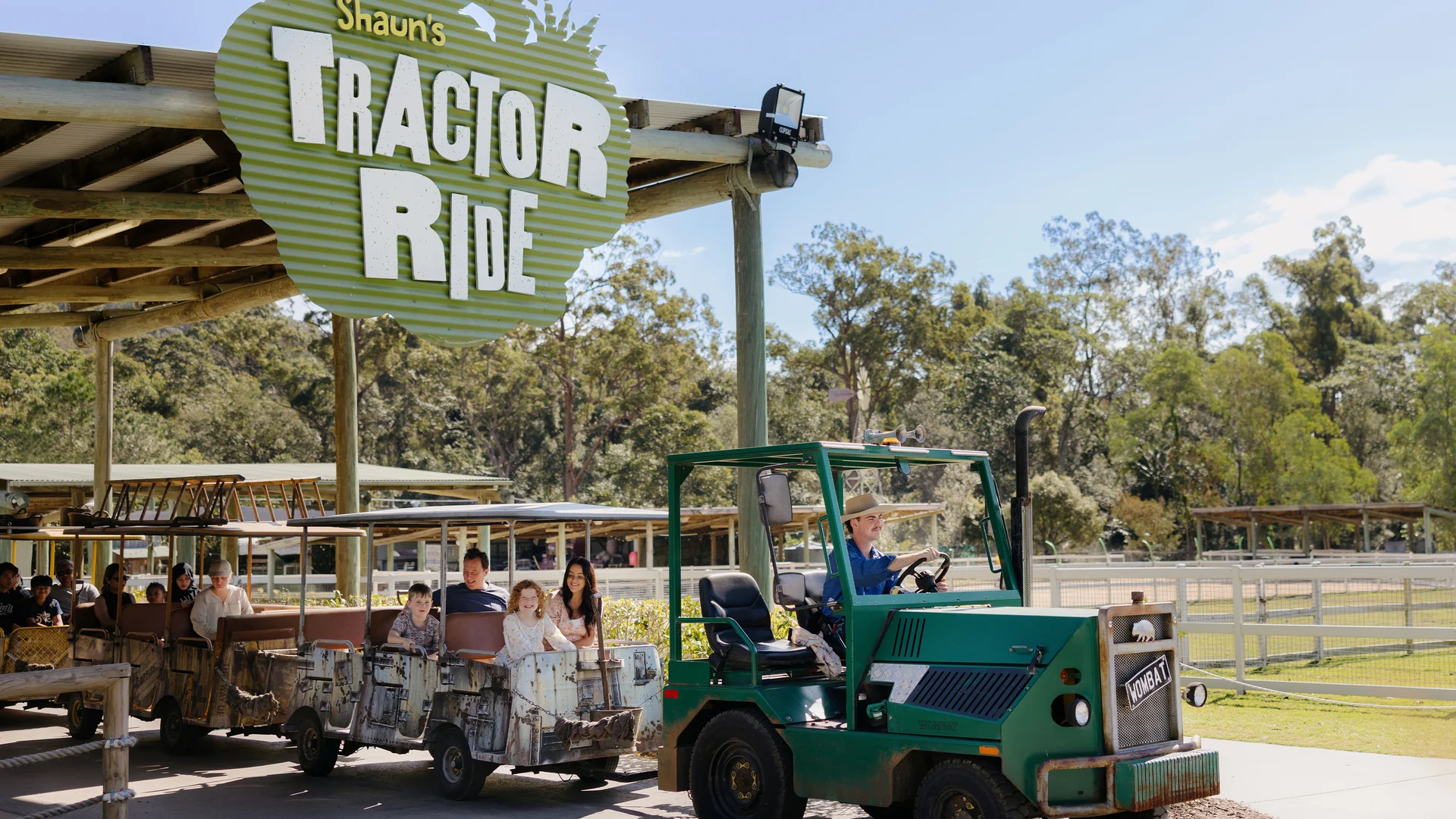 A green tractor pulls open-air carriages with seated passengers under a large sign that reads “Shaun’s Tractor Ride” at an outdoor farm attraction surrounded by trees and fences.