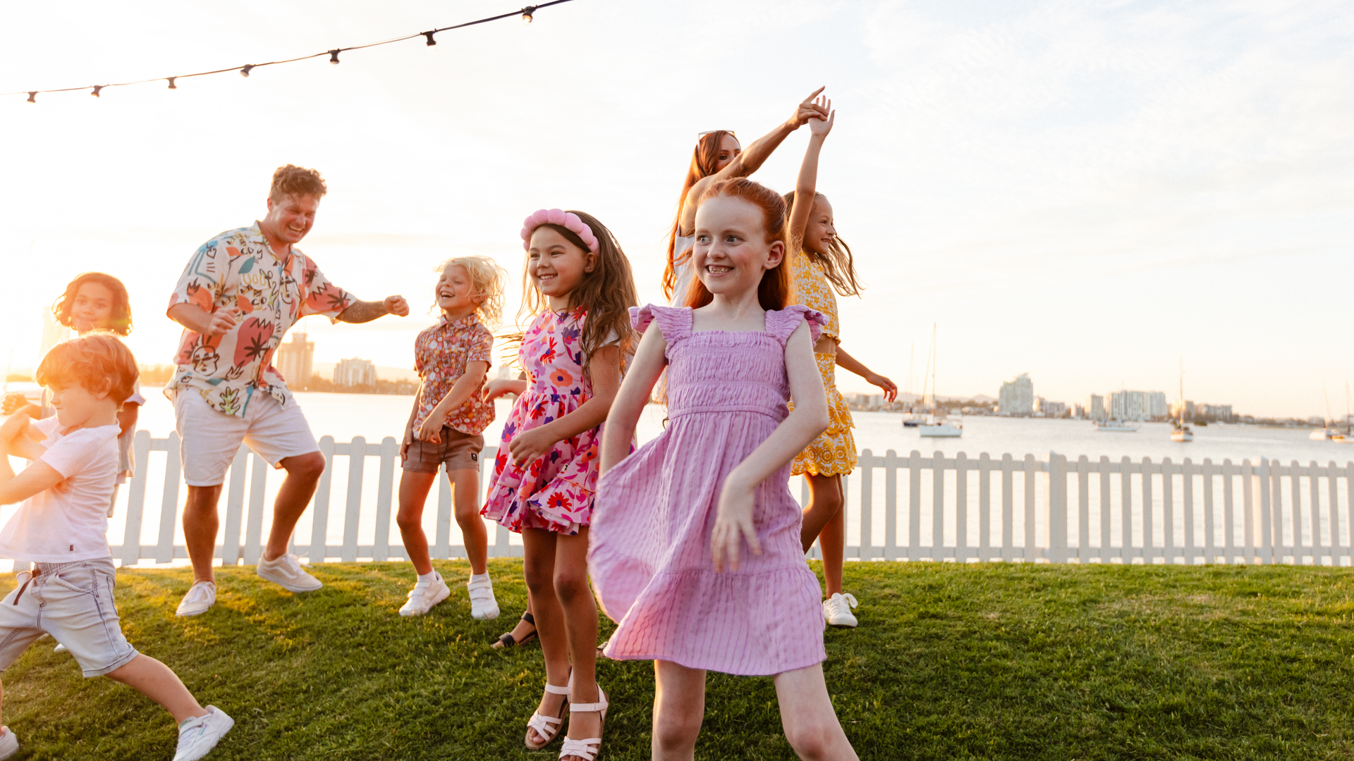 A group of smiling children and an adult dance and play on a grassy lawn near a white fence by the water at sunset, with city buildings and boats visible in the background.