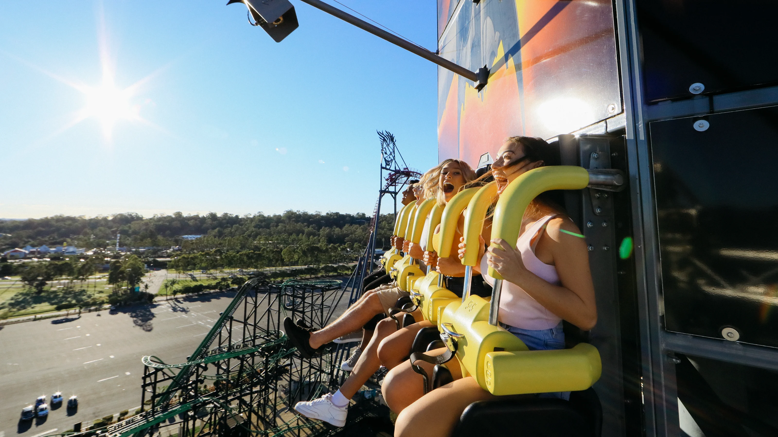 Three people are seated and secured in an amusement ride harness, elevated high above the ground on a sunny day, with a scenic view of the park and surroundings in the background.