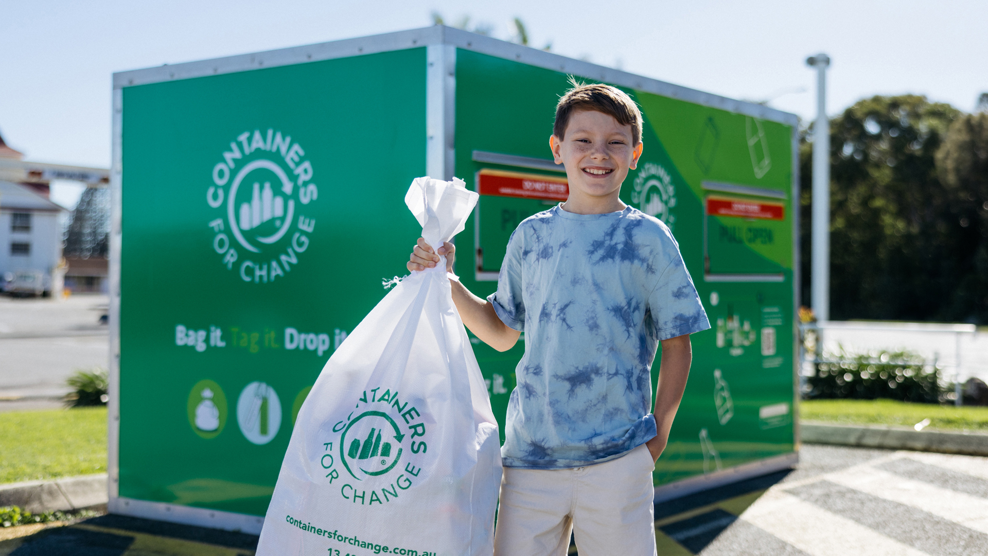 A boy holds a white recycling bag in front of a green "Containers for Change" collection bin at an outdoor recycling center.