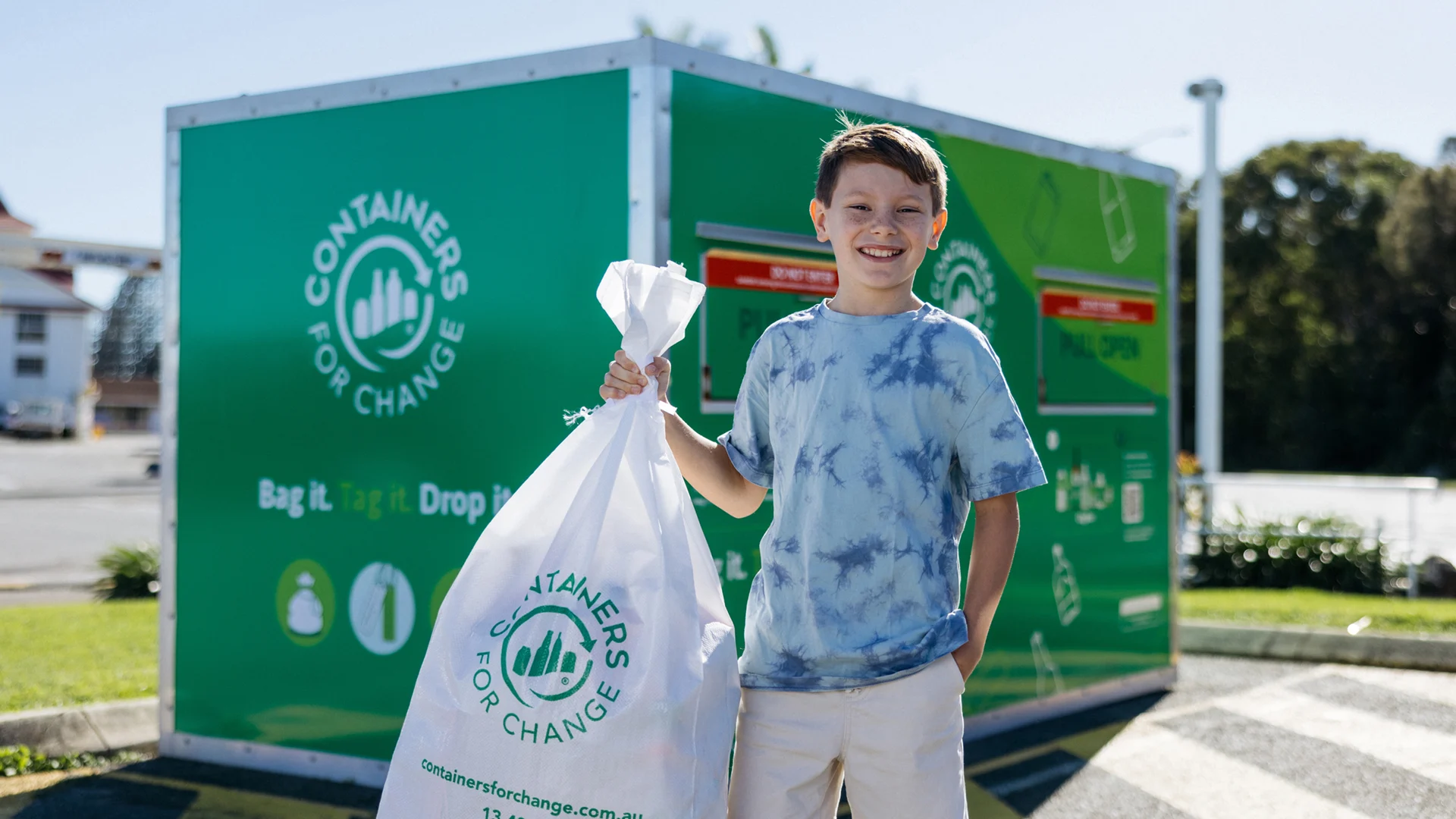 A boy holds a white recycling bag in front of a green "Containers for Change" collection bin at an outdoor recycling center.