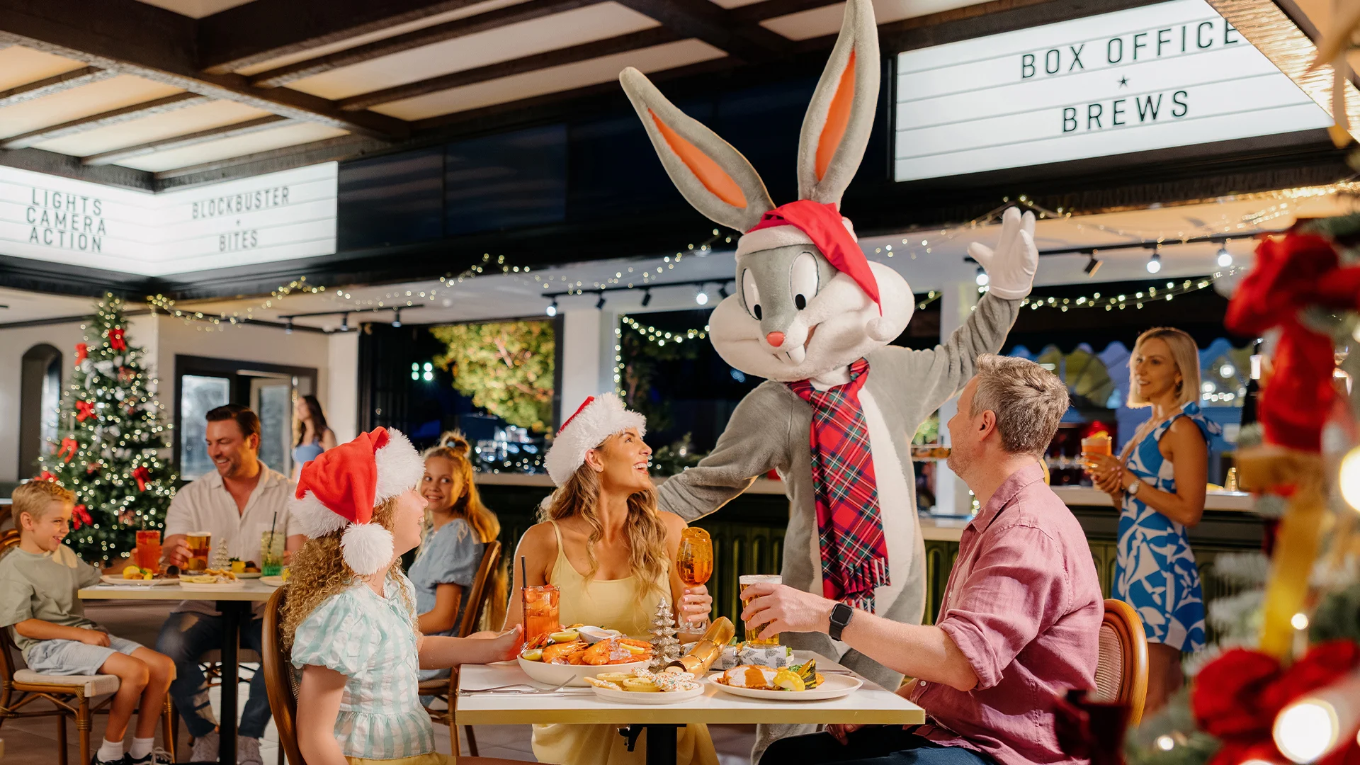 A family in festive attire dines at an outdoor restaurant decorated with Christmas lights and a tree. A person in a Bugs Bunny costume wearing a Santa hat and scarf greets them, while others enjoy food and drinks nearby.