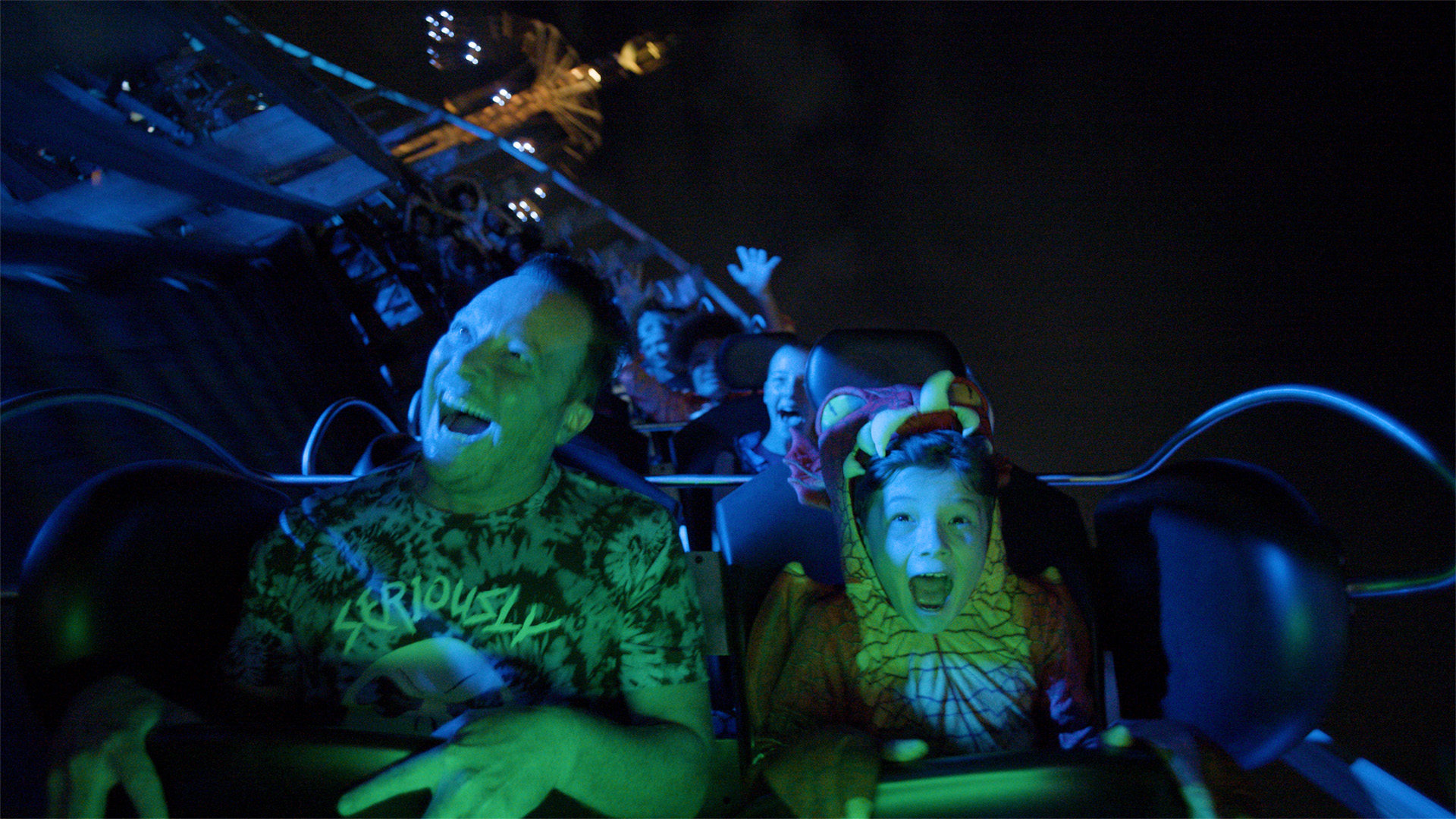 A group of people, including a child in a costume, ride a roller coaster at night, illuminated by blue lights. Their faces show excitement and fear as the ride speeds downward.