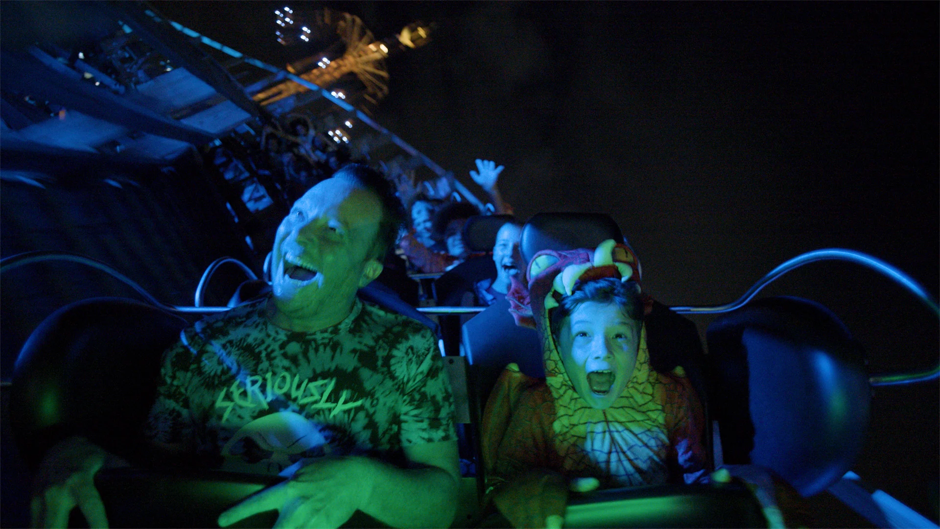 A group of people, including a child in a costume, ride a roller coaster at night, illuminated by blue lights. Their faces show excitement and fear as the ride speeds downward.