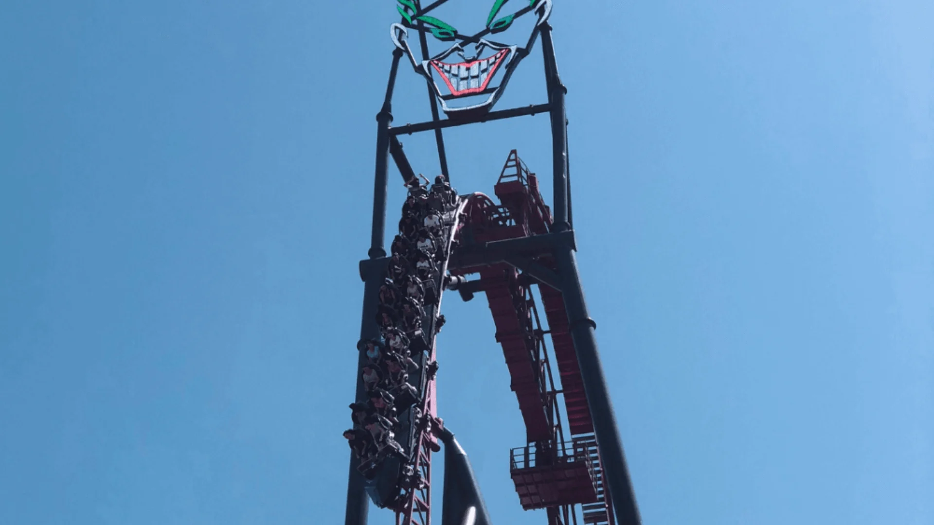 Riders plunging down the vertical drop of the DC Rivals HyperCoaster at Movie World, beneath the large illuminated Joker face sign against a clear blue sky.