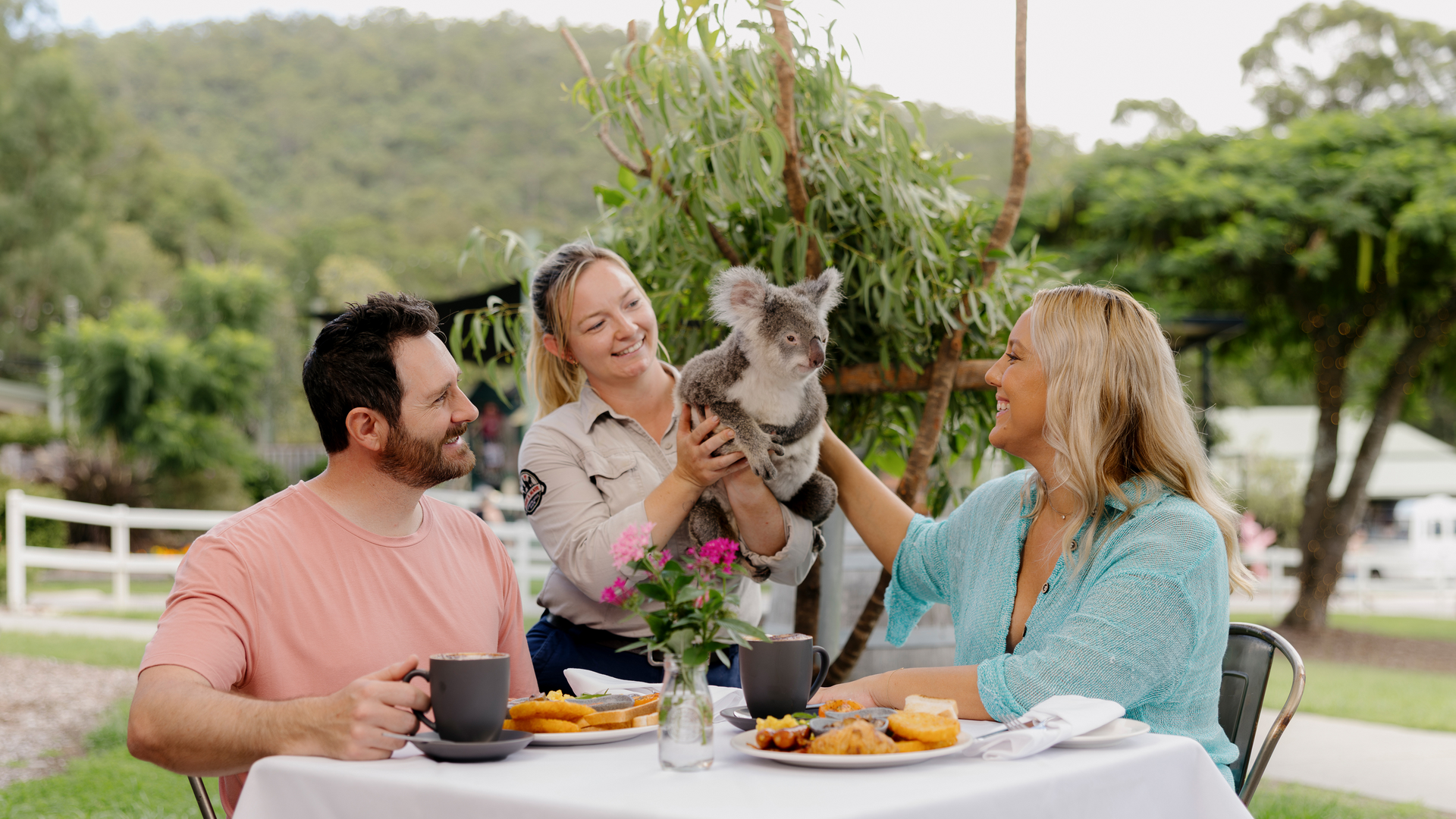 A smiling couple sits at an outdoor table enjoying breakfast while a wildlife handler presents a koala for them to pet. Trees and greenery surround the area, creating a peaceful setting.