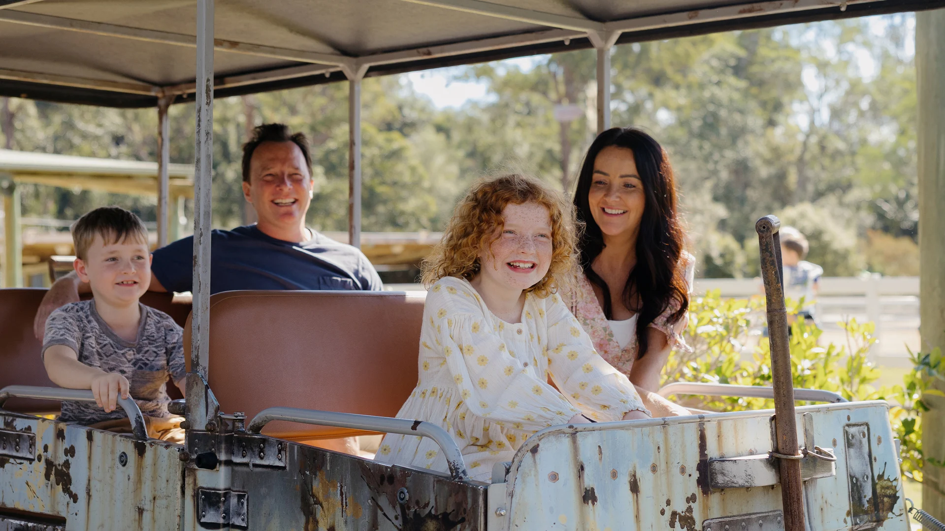 A family of four, two adults and two children, sit smiling on a rustic outdoor train ride in a sunny, natural setting with trees in the background.