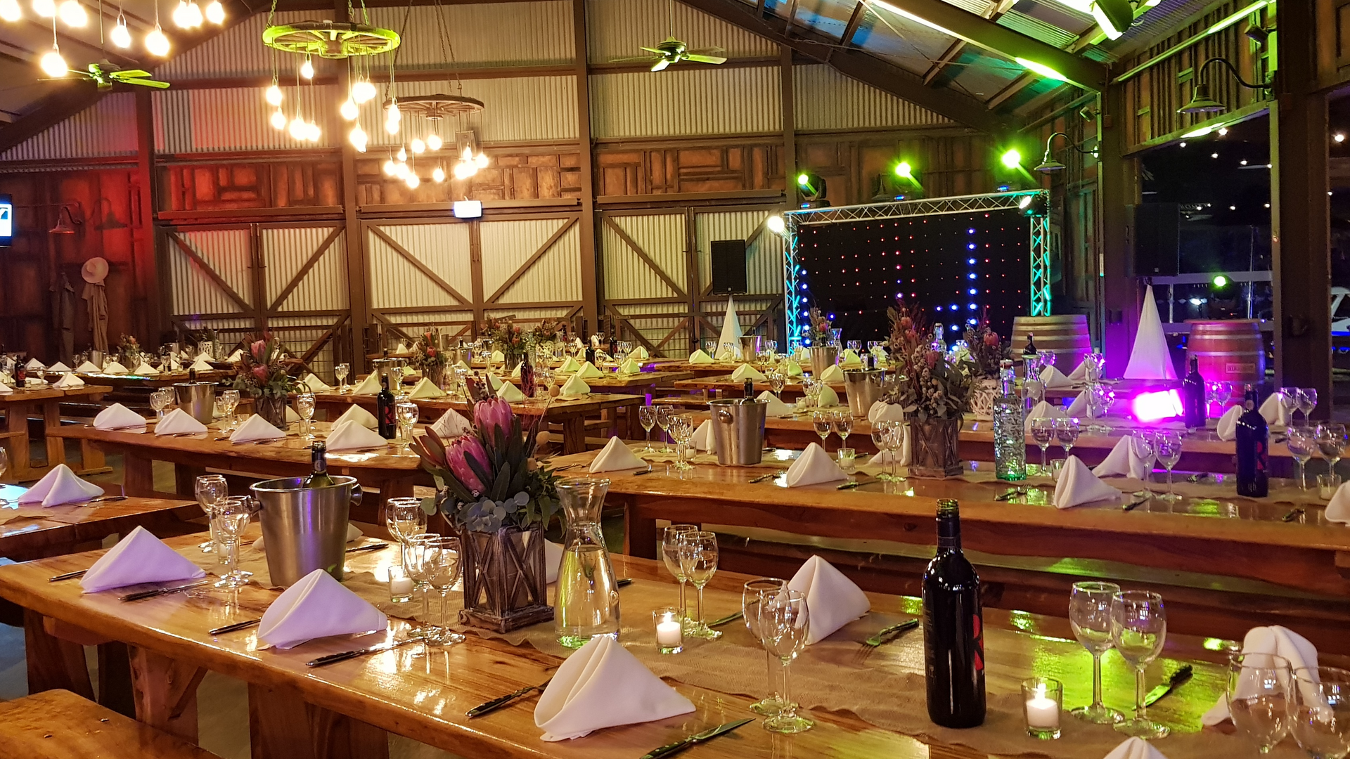 A large rustic banquet hall with wooden tables set for an event, decorated with flowers, candles, wine bottles, and folded napkins, with a lit stage in the background.
