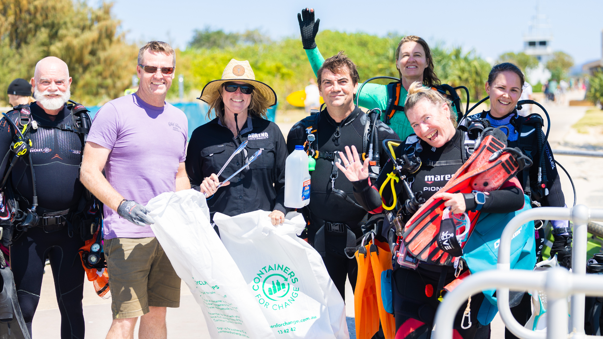A group of smiling people in wetsuits and casual clothes hold bags and diving gear, posing outdoors on a sunny day after a beach or ocean clean-up. Green trees and blue sky are visible in the background.
