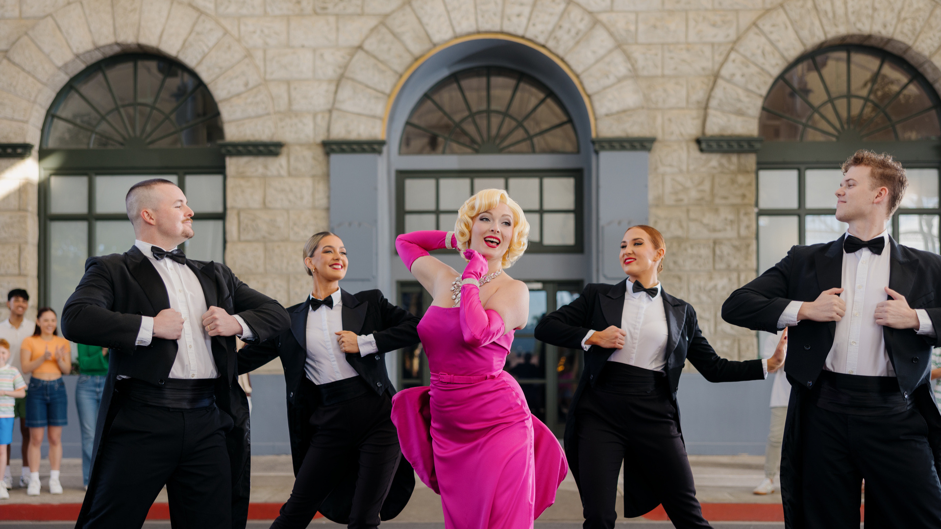 Marilyn Monroe 2024. A performer in a pink dress and four dancers in tuxedos pose in front of a building with arched windows.