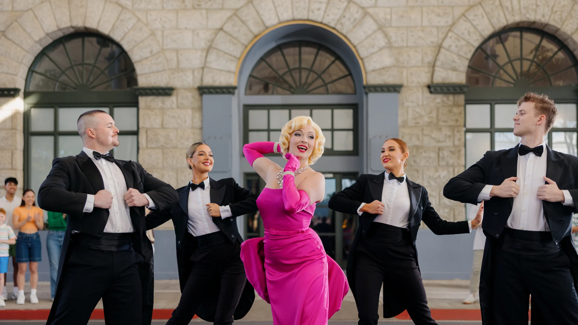 Marilyn Monroe 2024. A performer in a pink dress and four dancers in tuxedos pose in front of a building with arched windows.