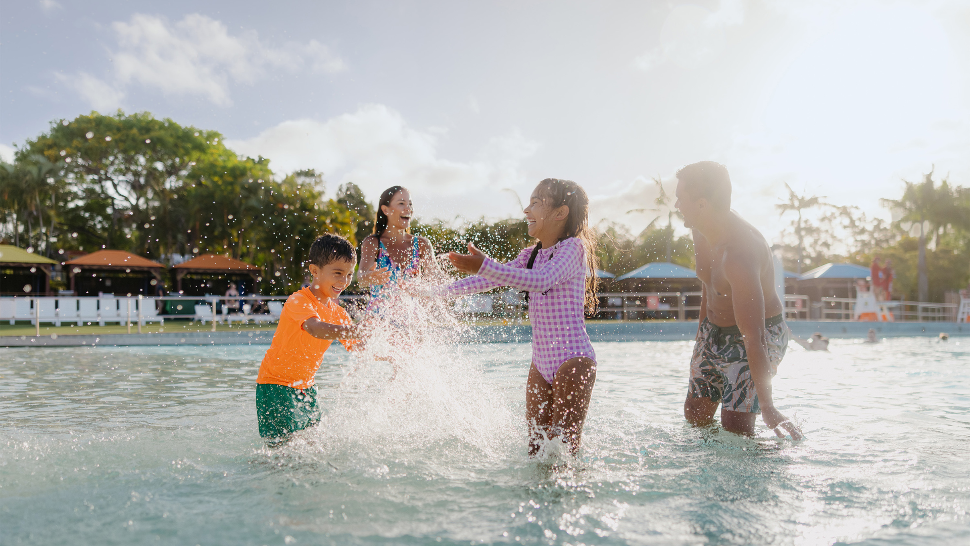 A family of four, two adults and two children, play and splash together in a shallow outdoor pool on a sunny day, surrounded by trees and colorful umbrellas in the background.