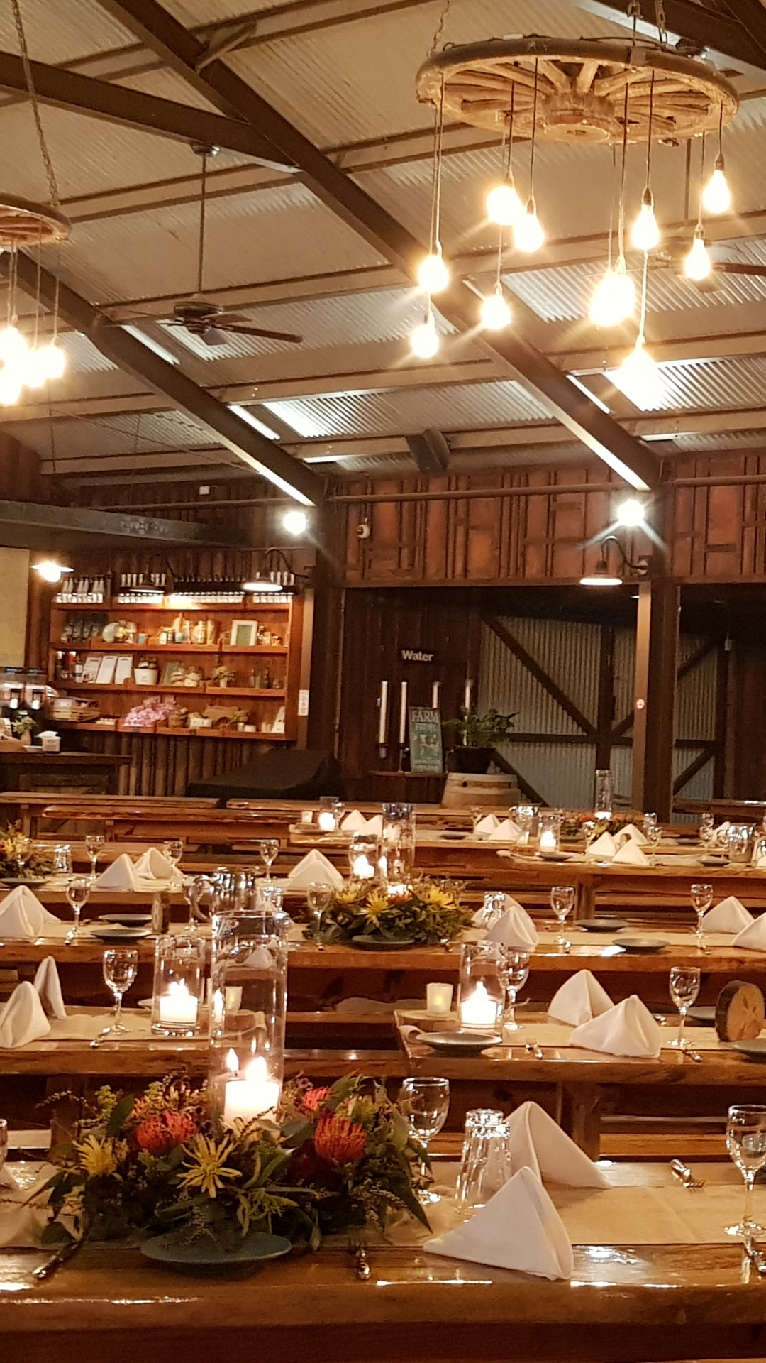 Large rustic dining hall with wooden tables set for a meal, decorated with flowers and candles, under warm hanging lights. Shelves and a counter are visible in the background.