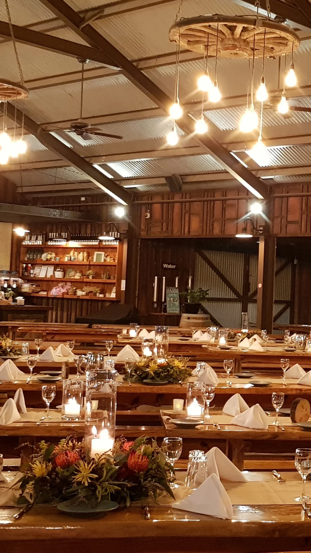 Large rustic dining hall with wooden tables set for a meal, decorated with flowers and candles, under warm hanging lights. Shelves and a counter are visible in the background.