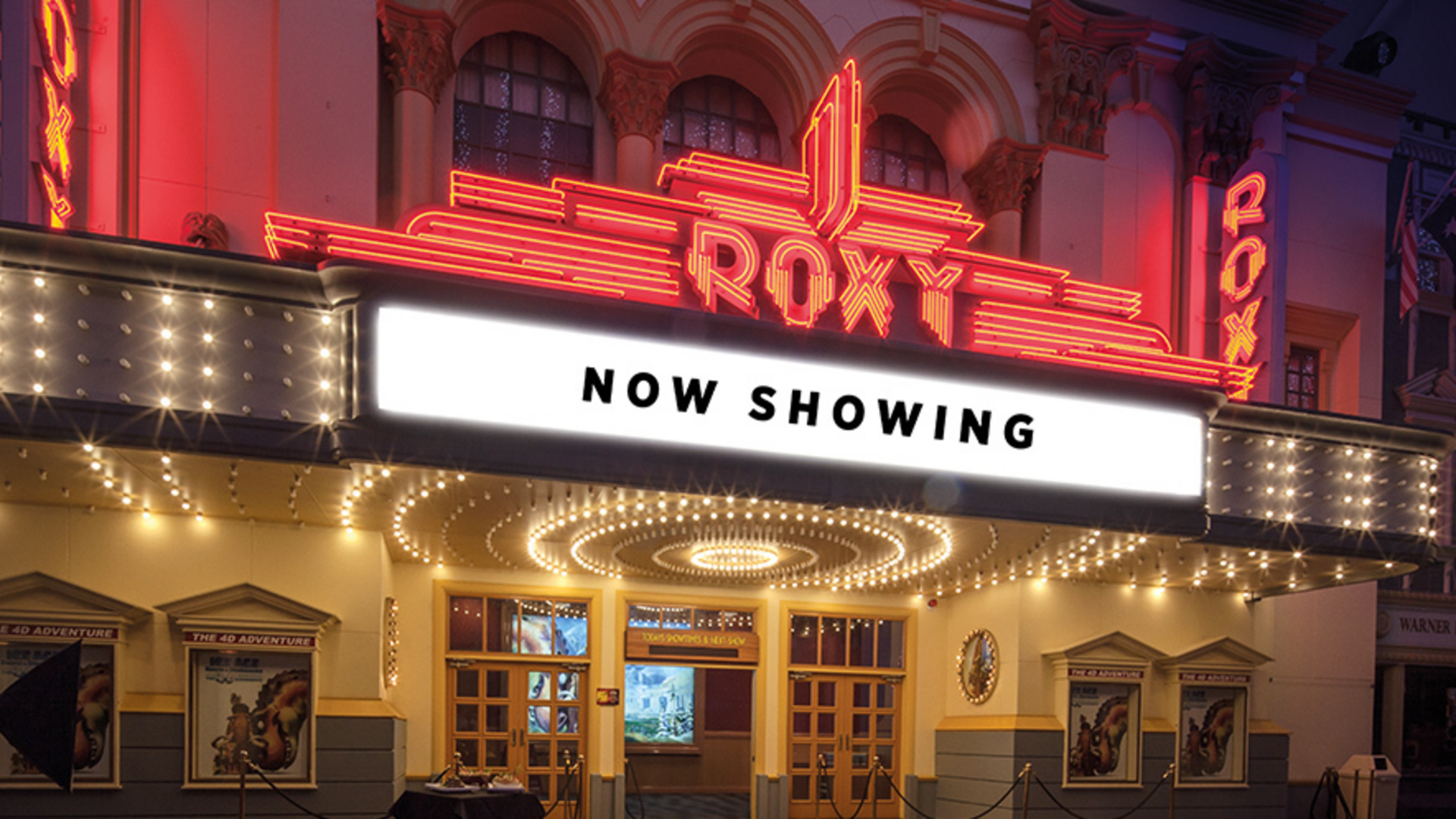 The brightly lit entrance of the Roxy Theater at night features a large marquee sign reading "NOW SHOWING" above the doors, with neon lights and ornate architectural details.
