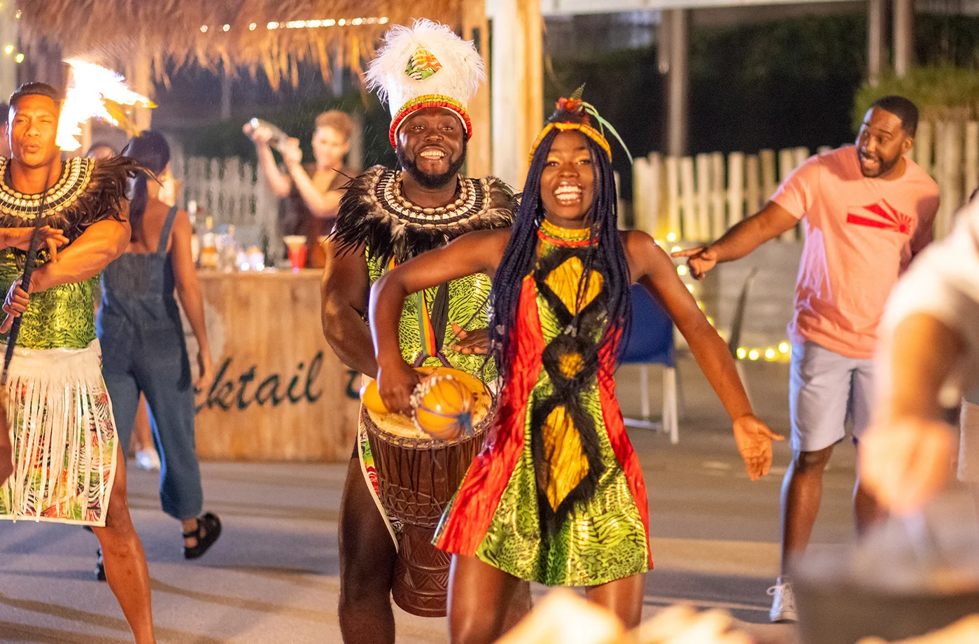 A man and woman in colorful, traditional costumes perform a lively dance and drum at an outdoor event, while people watch and take photos in the background. String lights and a tiki-style bar are visible.