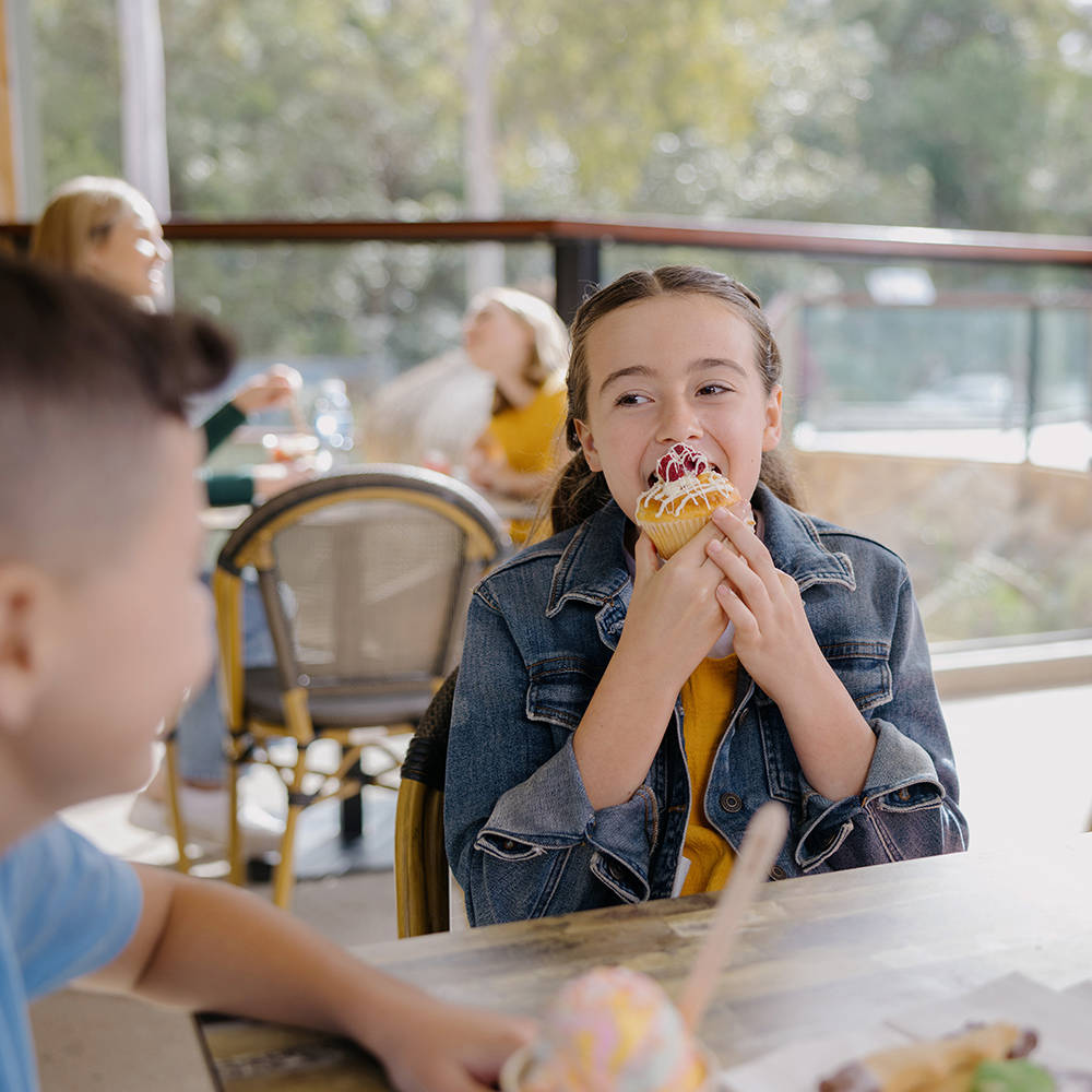 A young girl in a denim jacket eats a cupcake with sprinkles at a table, while a boy sits across from her with ice cream in front of him.