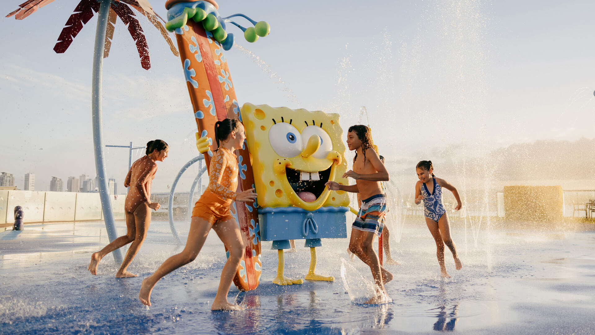 Children play around a SpongeBob SquarePants water feature in an outdoor water park, with sprays of water around them.
