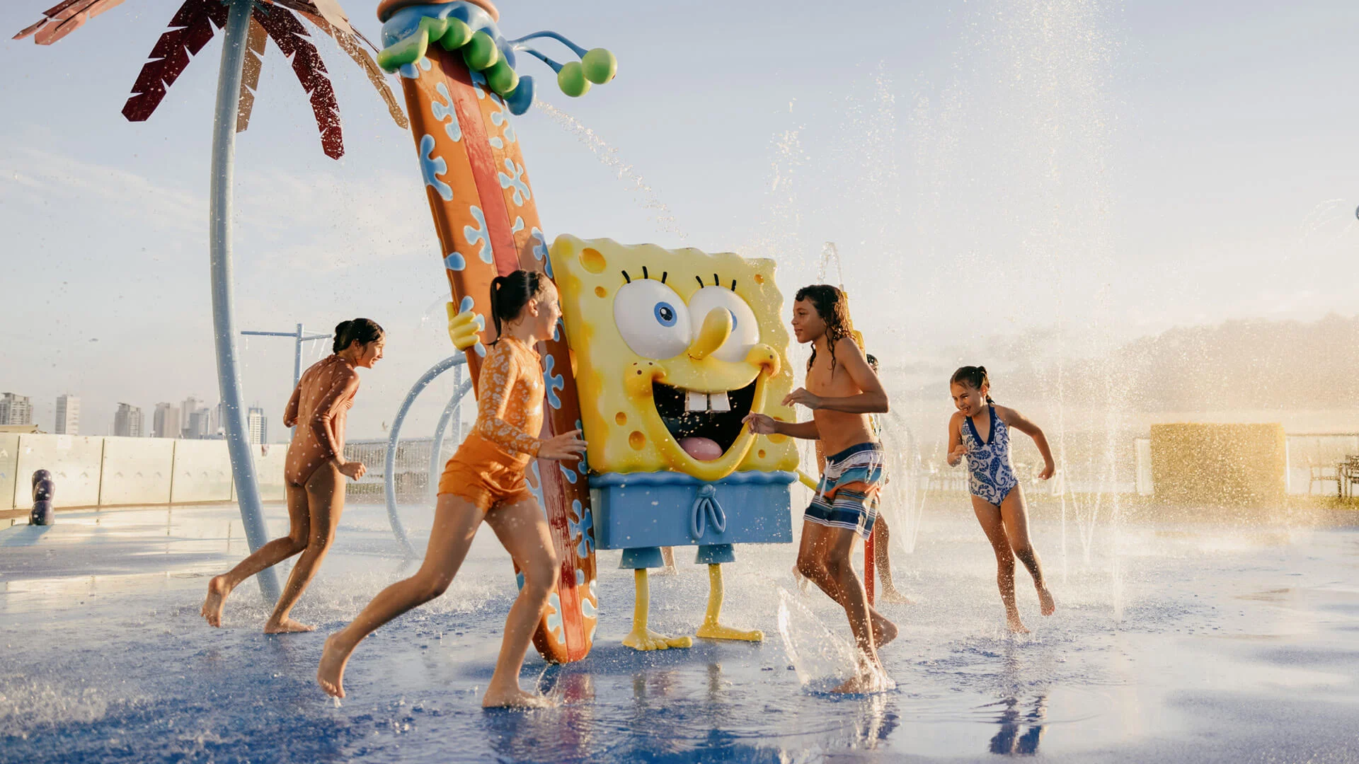 Children play around a SpongeBob SquarePants water feature in an outdoor water park, with sprays of water around them.
