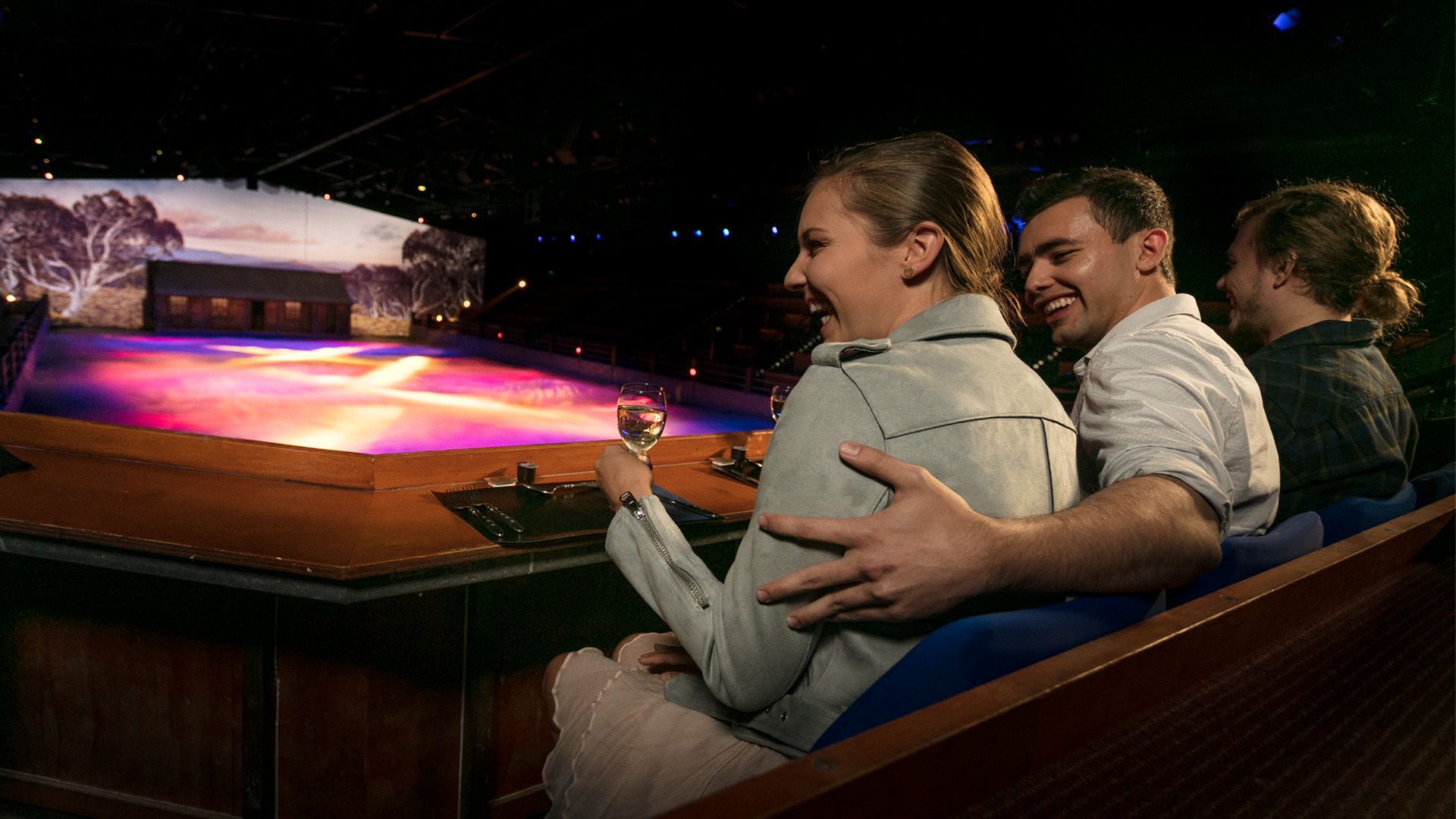 A smiling couple sits in an arena, enjoying drinks and watching a brightly lit stage show with colorful lights and scenery, while another person sits beside them.